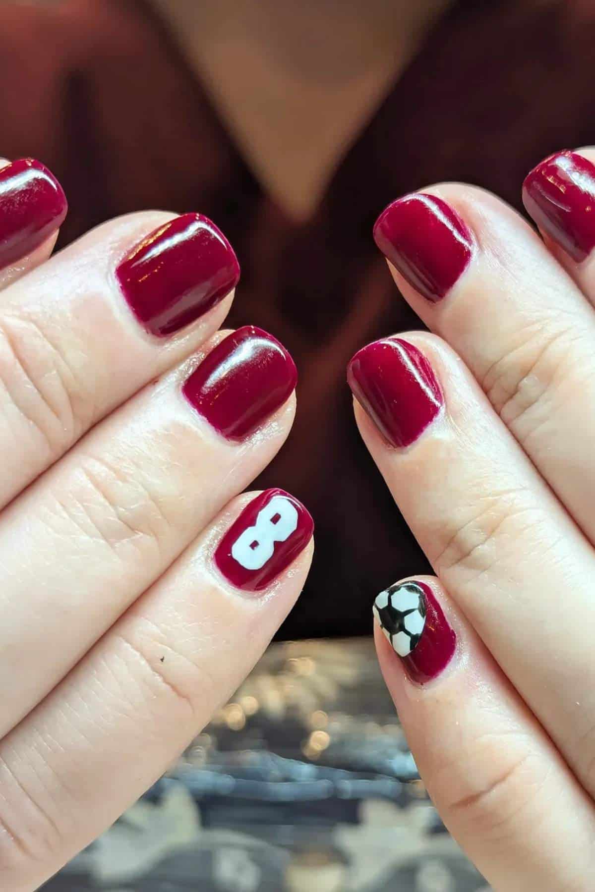Close-up of hands with short, dark red manicured nails; one nail has a white number 8, and another has a black and white soccer ball design.