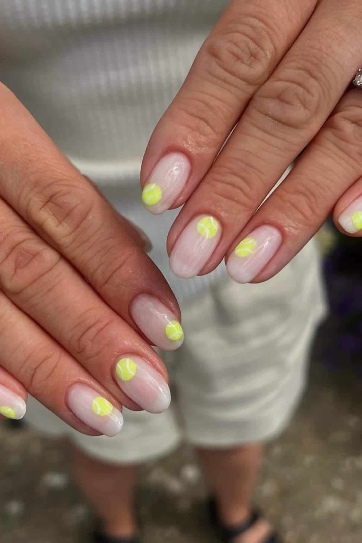 Close-up of hands with manicured nails, featuring a light pink base and small yellow tennis ball designs on each nail.