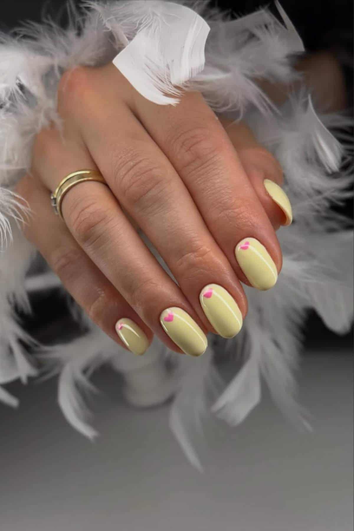 A hand with pale yellow oval-shaped nails, each featuring a small pink heart near the cuticle, surrounded by white feathers.