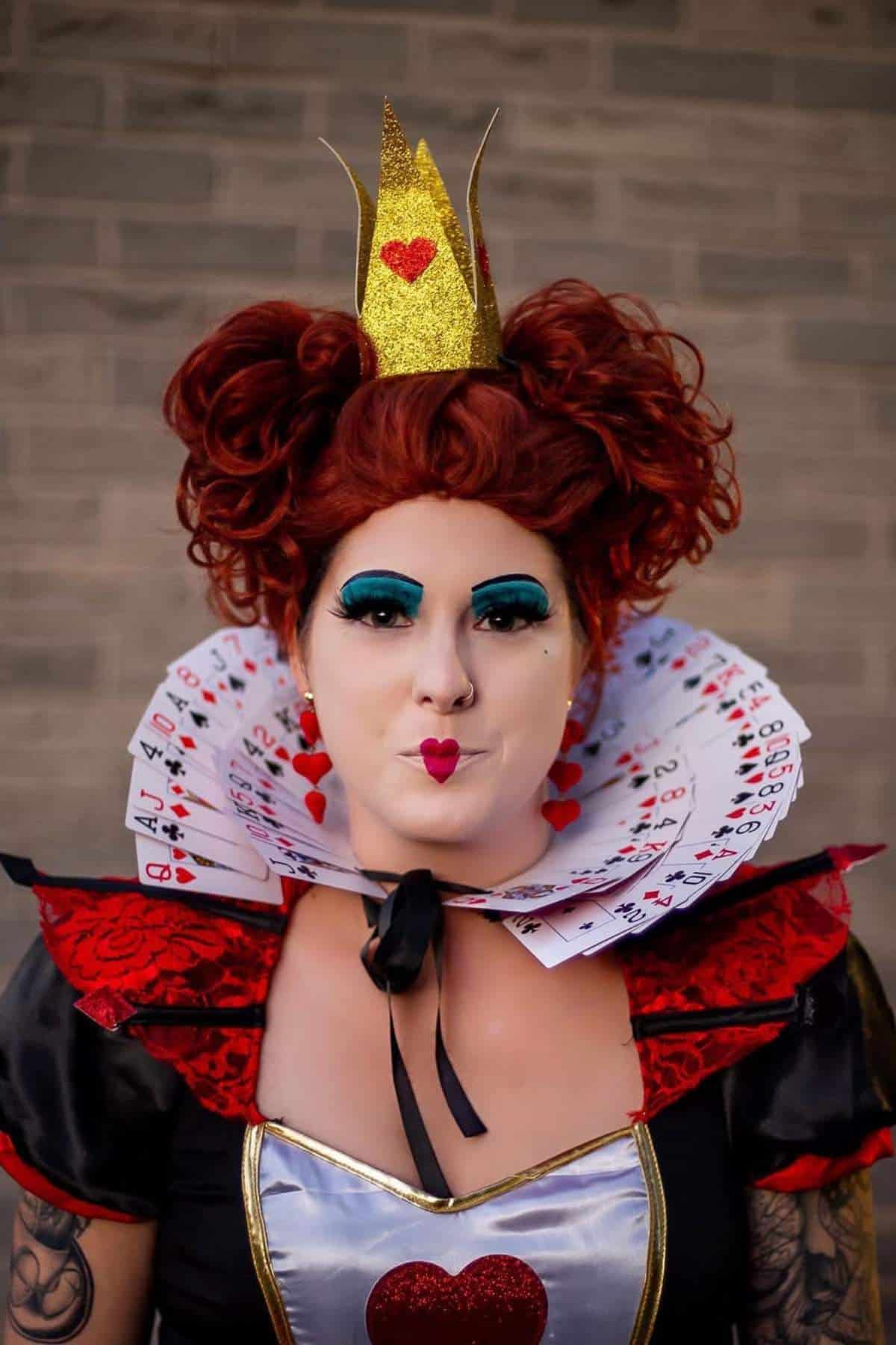 Person dressed as the Queen of Hearts with a golden crown, bold makeup, and a collar made of playing cards against a brick wall background.