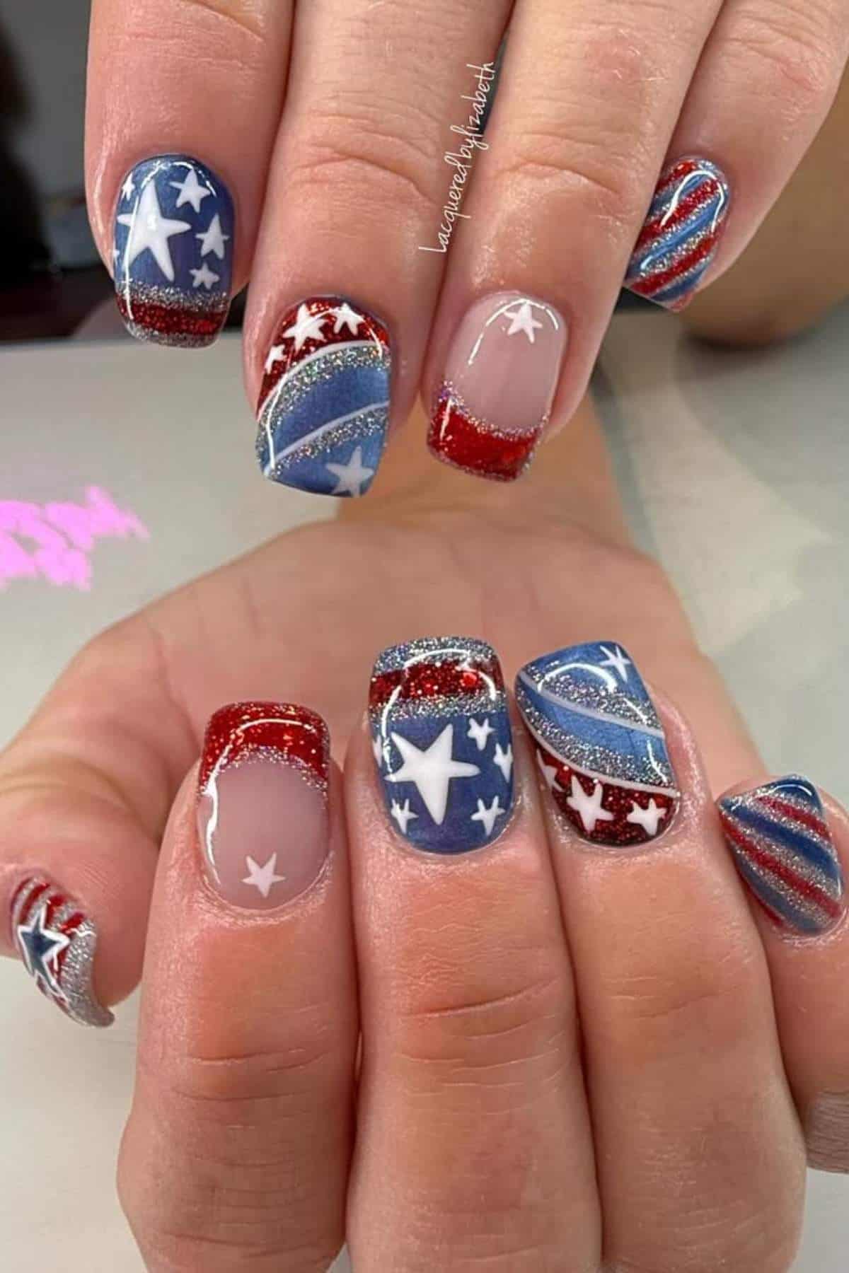 Close-up of hands with nails painted in red, white, and blue patriotic designs featuring stars, stripes, and glitter accents.
