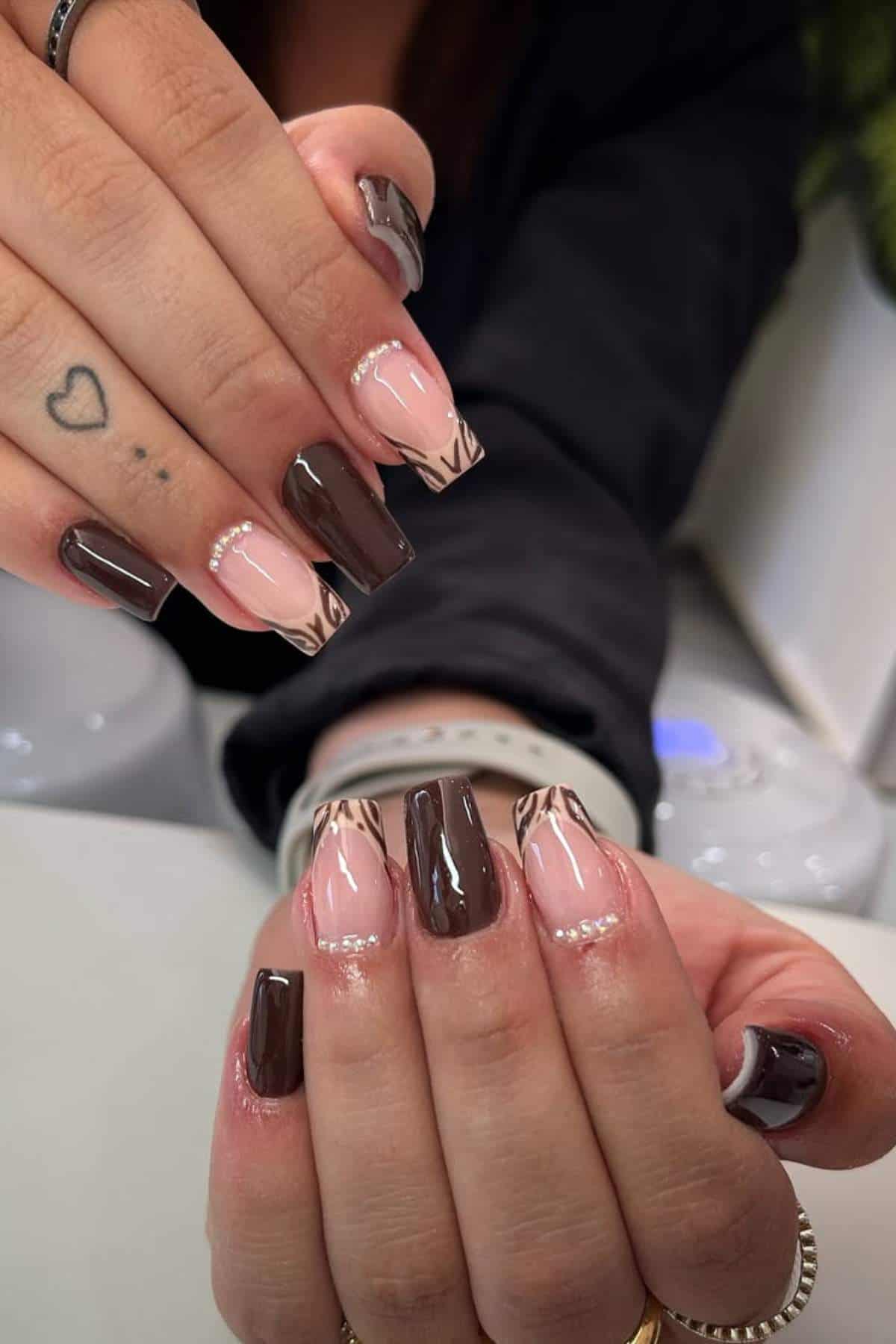 Close-up of hands with manicured nails featuring alternating glossy brown polish and nude polish with brown abstract tips, accented by a thin silver line near the cuticle.