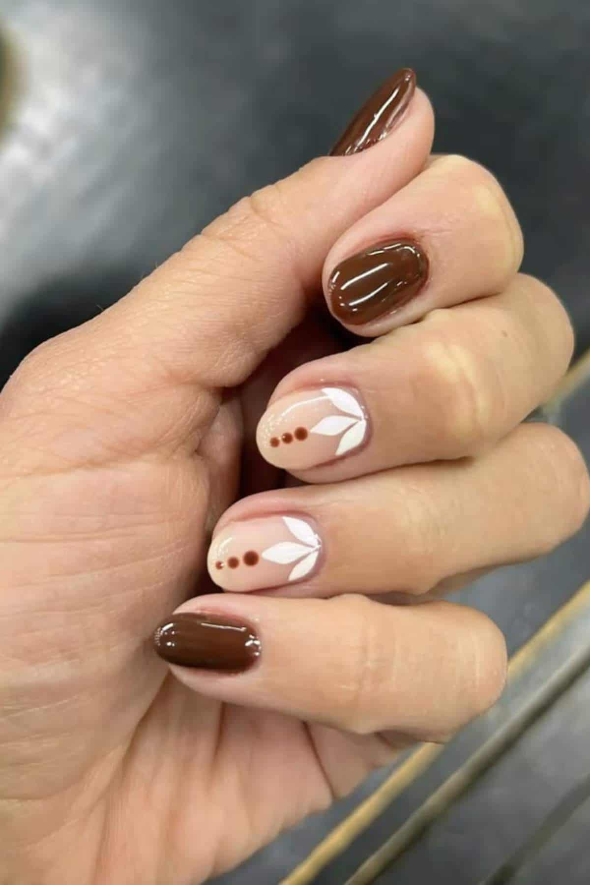A hand with manicured nails; most nails are painted brown, while two feature beige polish with white leaf designs and brown dots as accents.