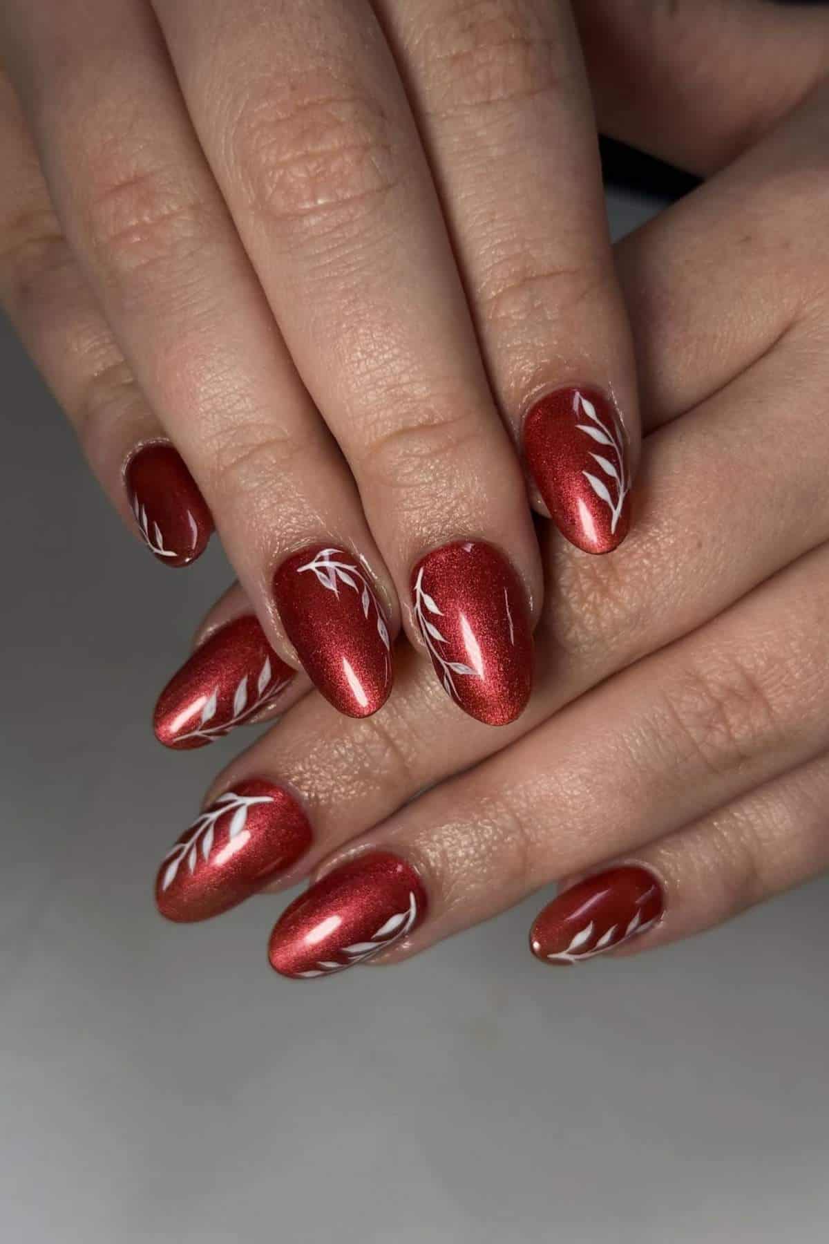 Hands with almond-shaped, metallic red nails featuring white leaf designs on some nails.