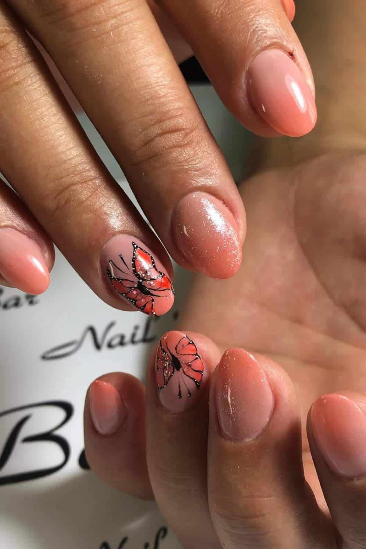 Close-up of hands with glossy peach-colored nails; two nails feature butterfly designs with orange and black detailing.