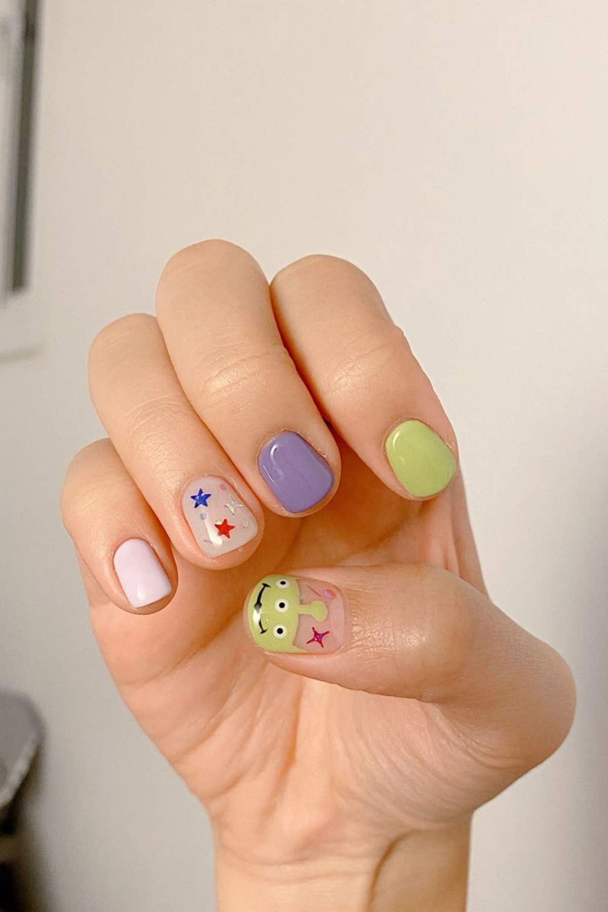 A hand with short, manicured nails painted in pastel colors with colorful star stickers and a cartoon frog design on the thumb.