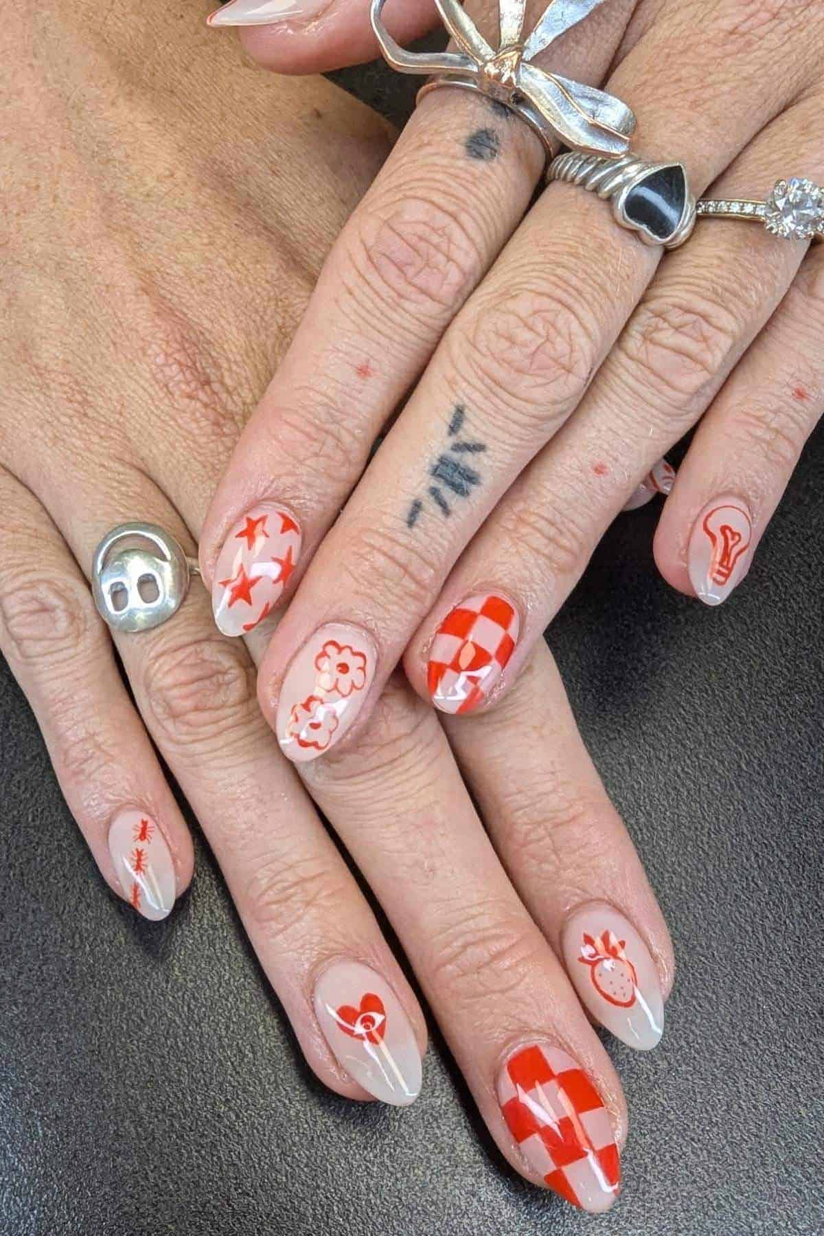 Hands with several silver rings and tattoos are shown; fingernails are painted with red and off-white designs, including stars, flowers, checkerboards, and abstract shapes.