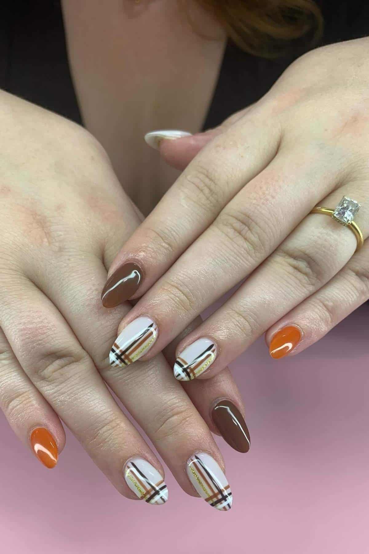 Hands with almond-shaped nails featuring a mix of brown, orange, and plaid designs. One hand wears a gold ring with a square-cut stone.