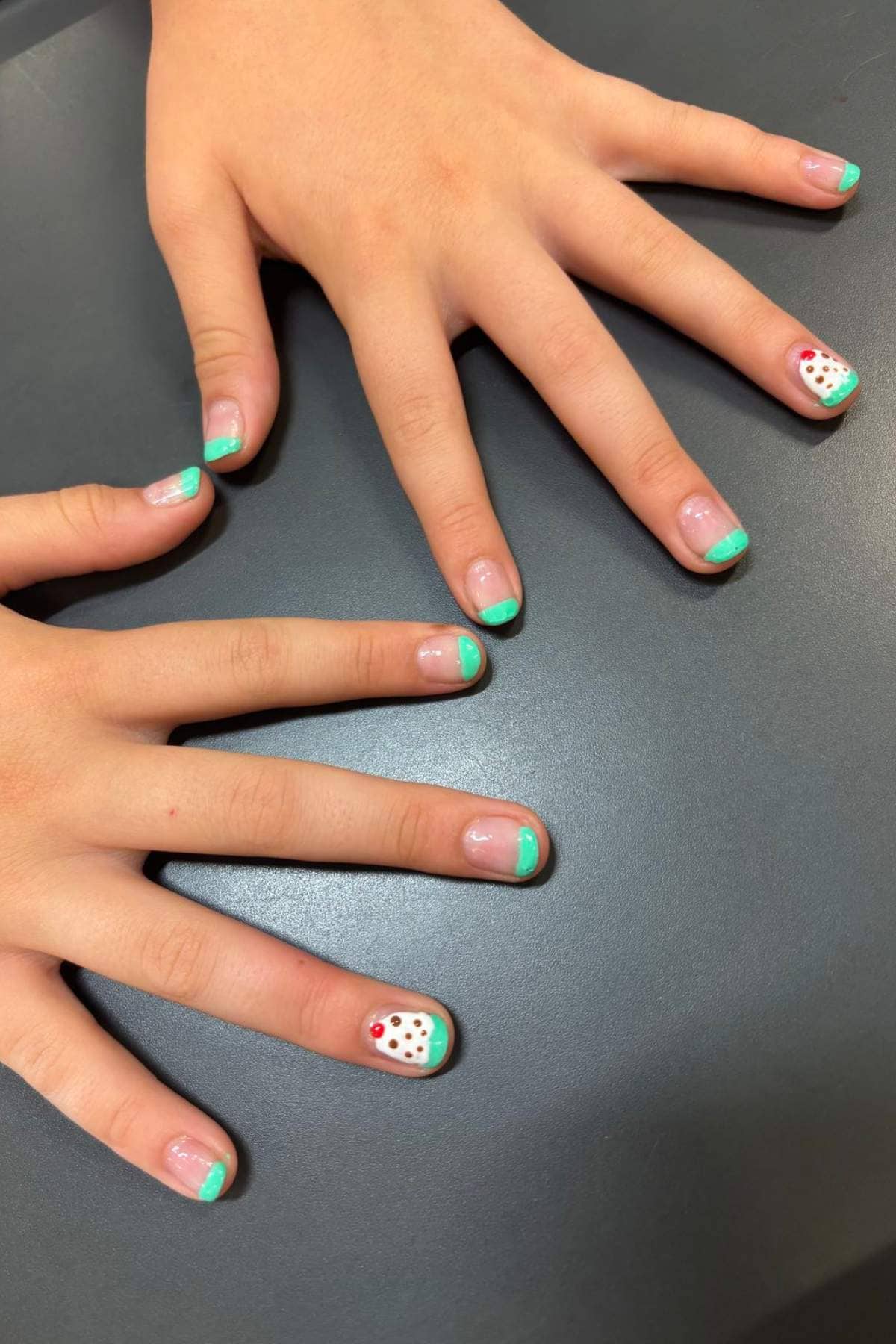 Two hands with nails painted with turquoise tips; the ring fingernails feature a white base with red and green polka dots. Hands are placed on a dark surface.