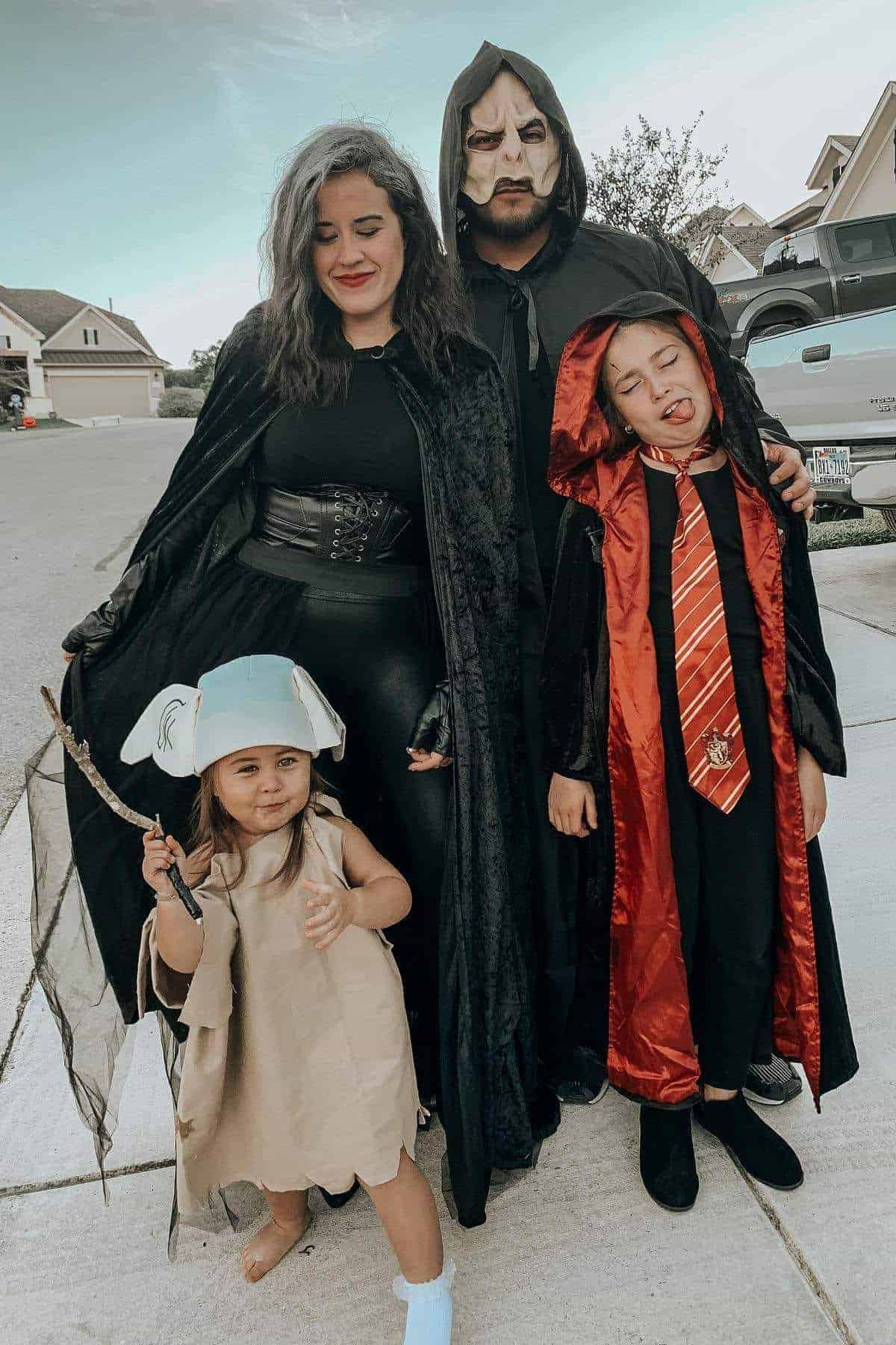 A family dressed in Harry Potter-themed costumes stands outside on a residential street, including a child as Dobby and others in wizard robes and cloaks.
