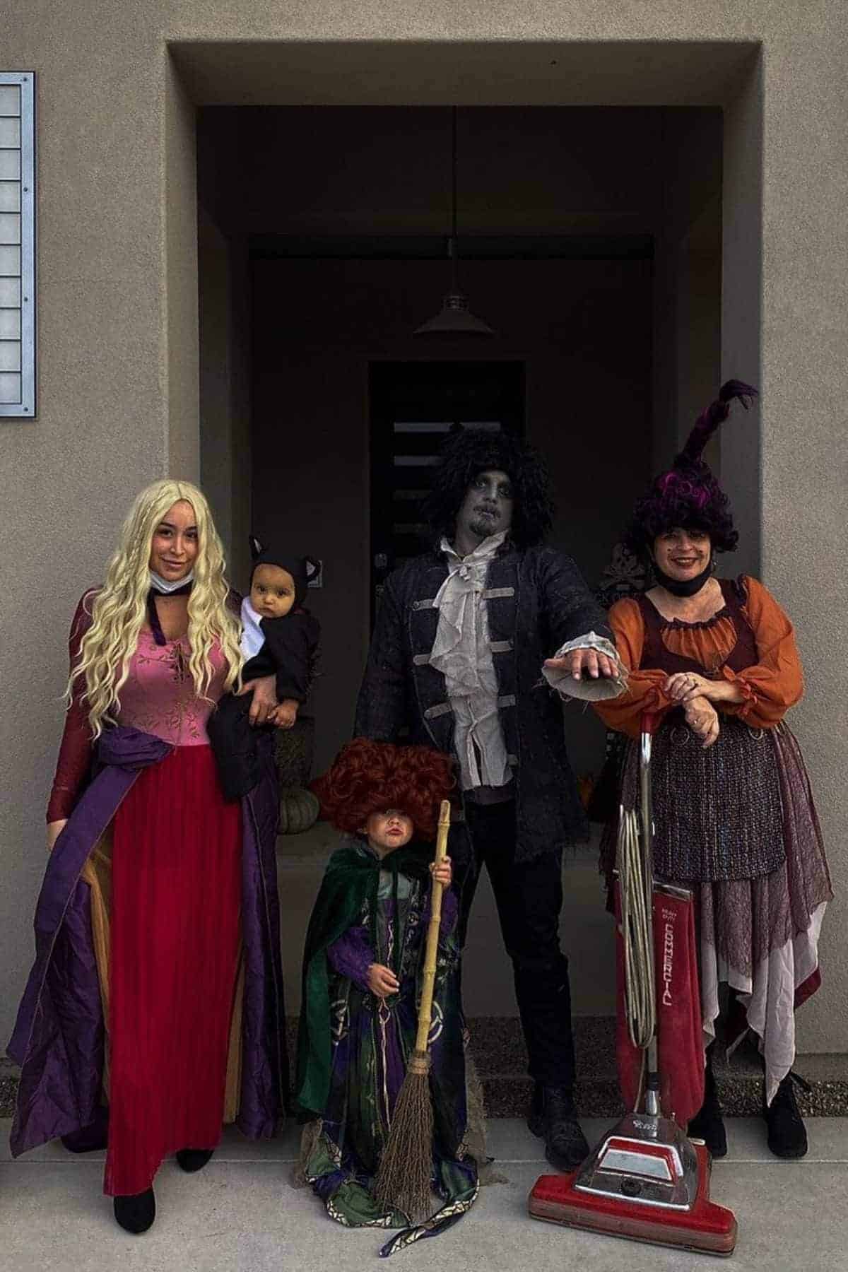 Five people in Halloween costumes pose outside a house, dressed as various characters, including witches and a vampire, with one child holding a broom and another adult with a vacuum cleaner.