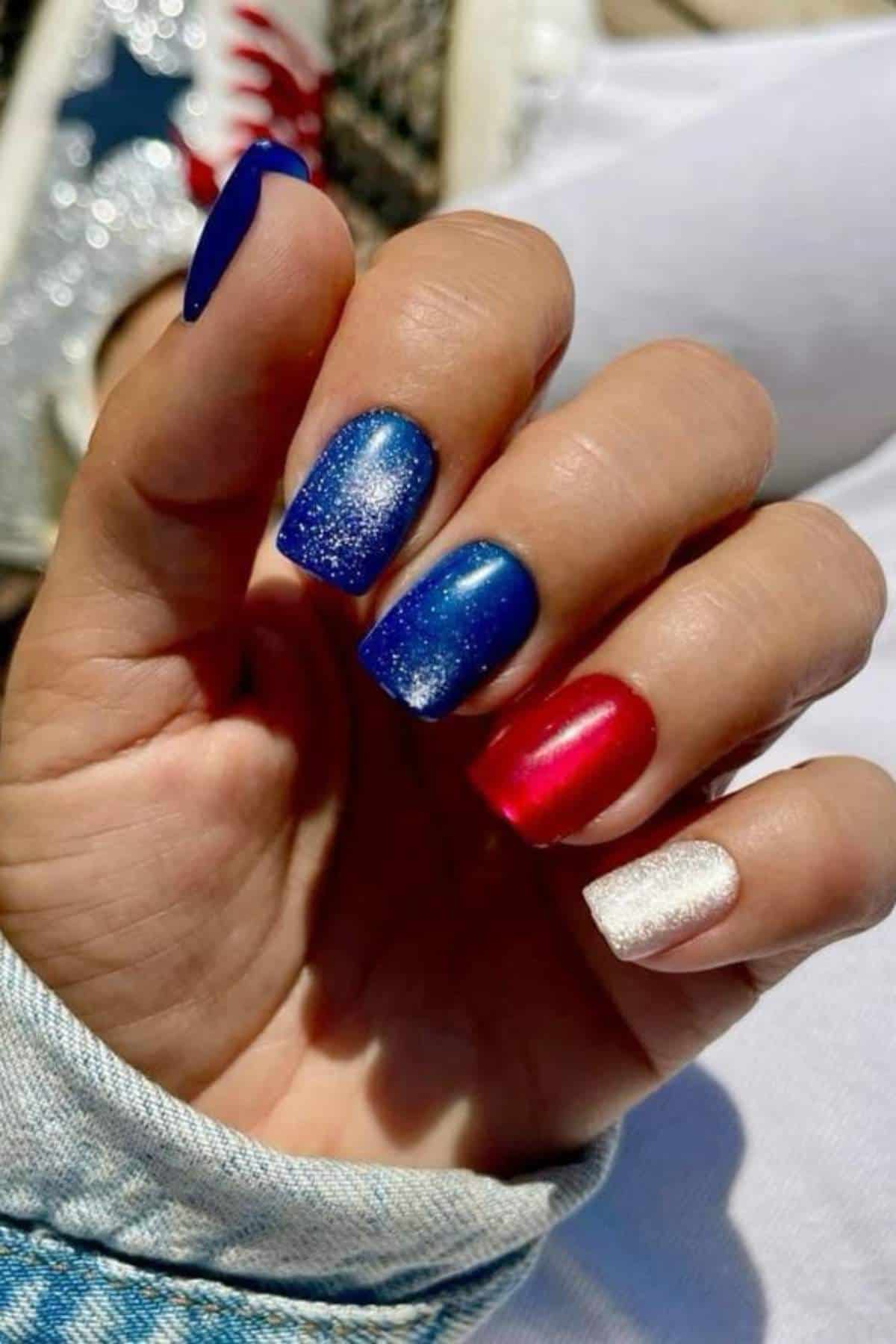 A hand with manicured nails painted in blue, glittery blue, red, and white colors, holding an object with a denim sleeve visible.