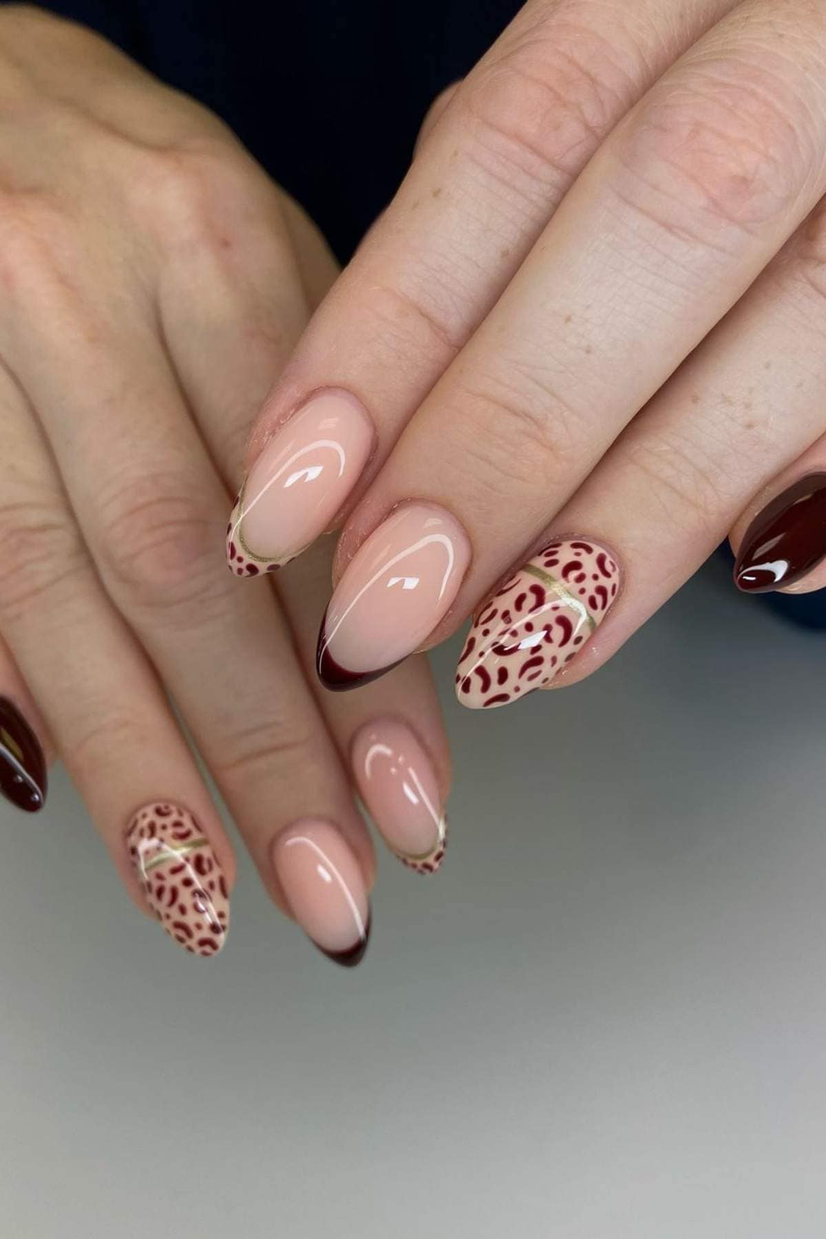 A hand with almond-shaped nails featuring burgundy French tips, burgundy animal print accents, and solid burgundy nail polish.