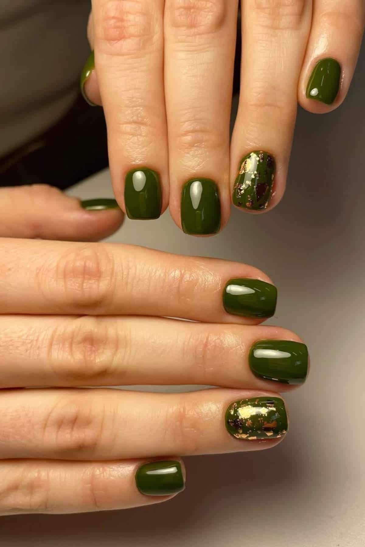 Close-up of hands with short, square nails painted dark green; one accent nail on each hand features gold and green foil decoration.