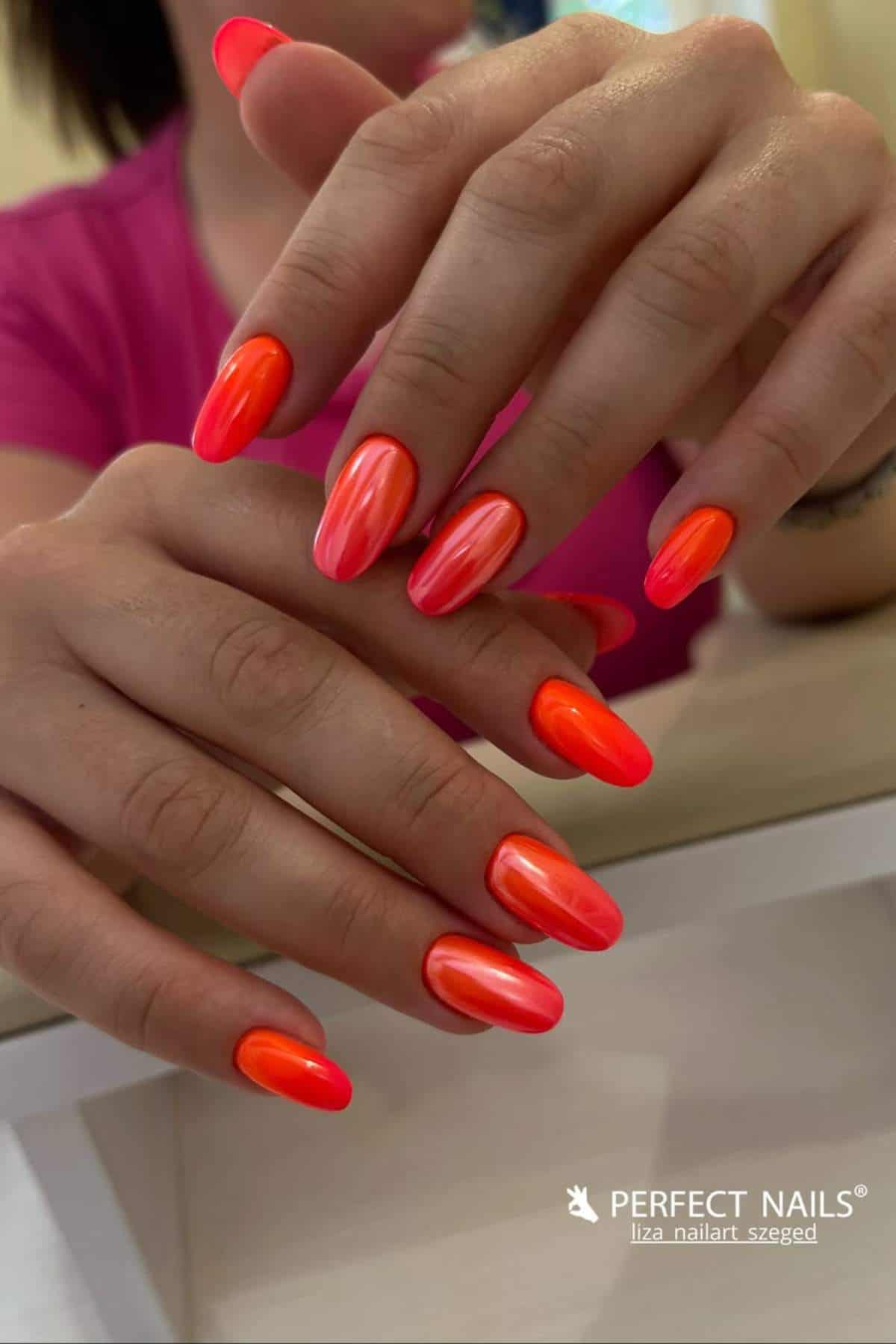 A person with long, almond-shaped nails painted in glossy shades of bright pink and orange, holding their hands together over a white surface.