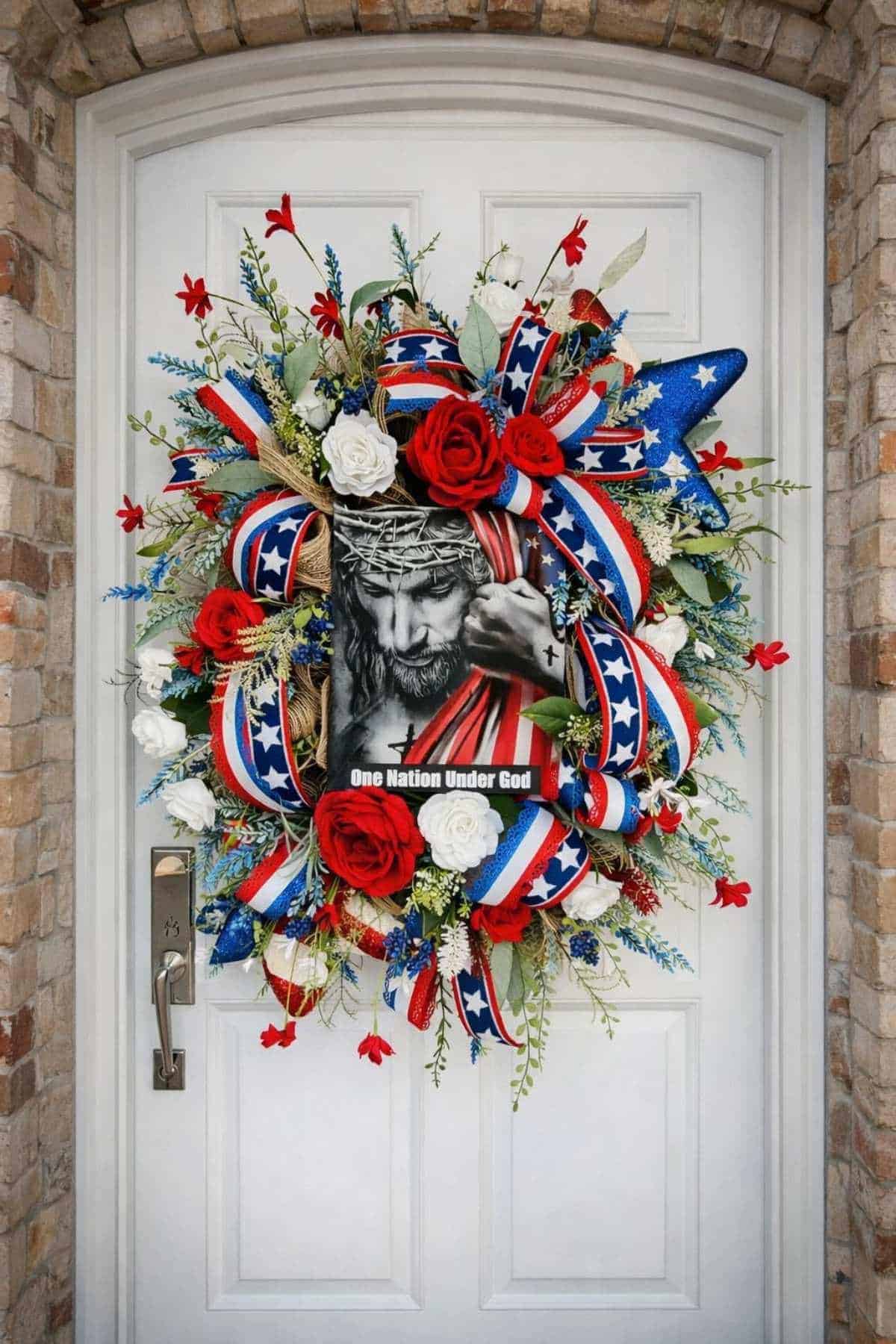 A patriotic wreath with red, white, and blue ribbons, artificial flowers, and an image of Jesus with the text "One Nation Under God" hangs on a white front door.