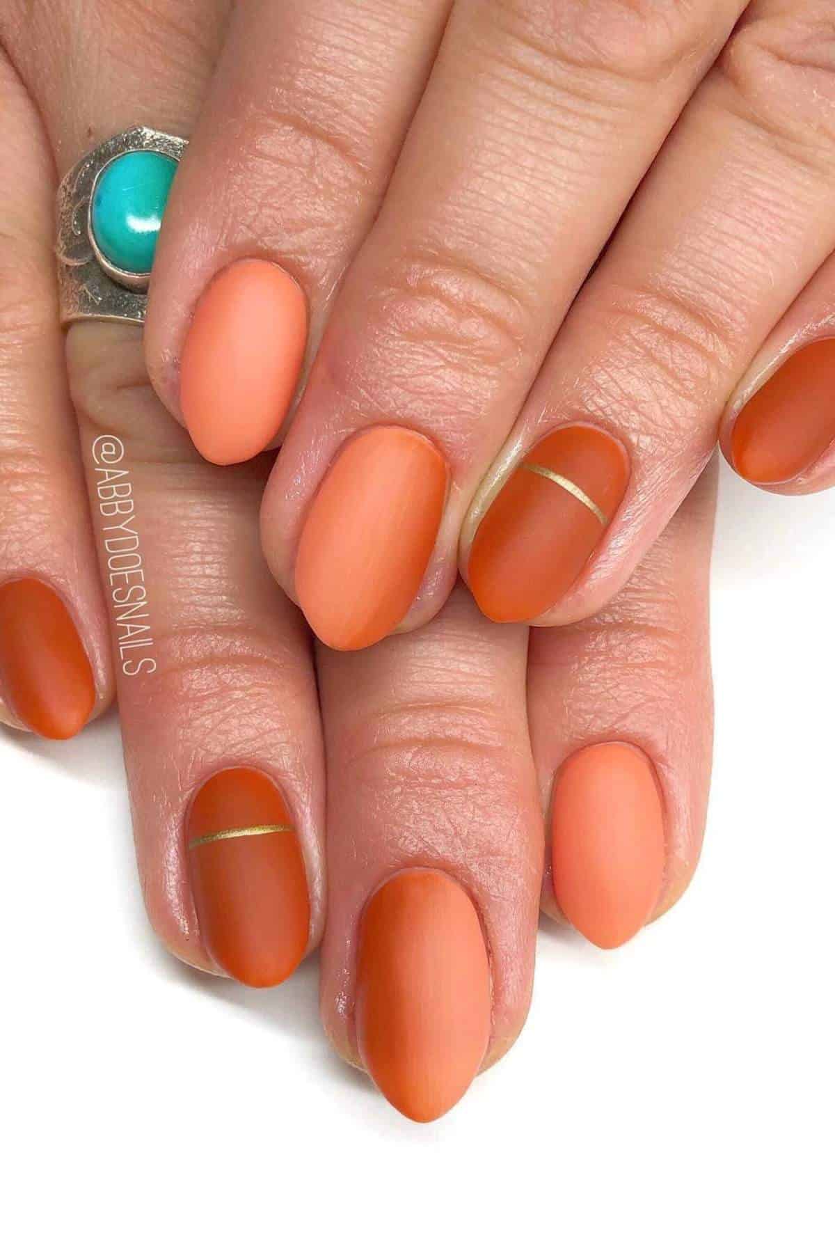 Close-up of hands with almond-shaped nails painted in matte orange and terracotta shades, some nails featuring a thin metallic gold stripe as a design detail.