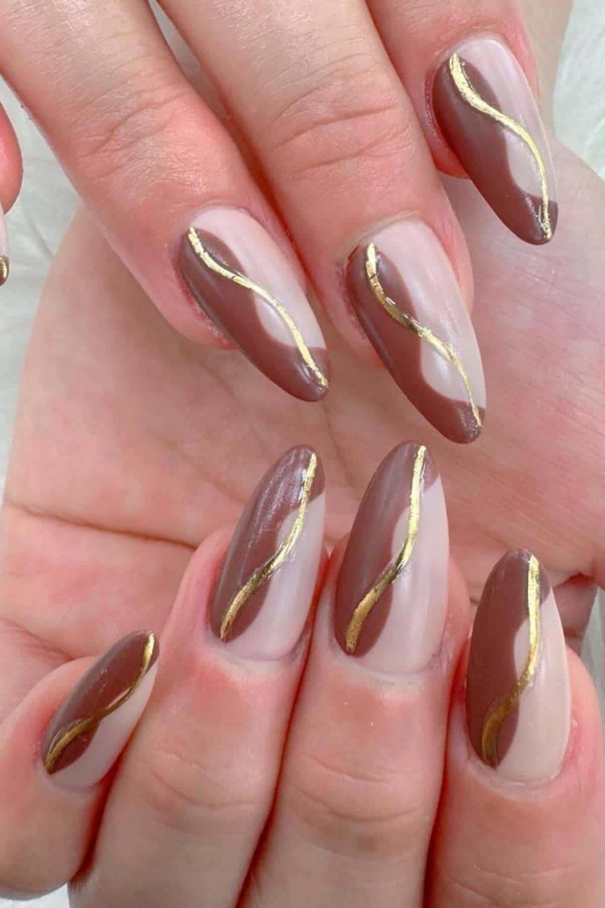 Close-up of hands with almond-shaped nails, painted in brown and nude tones with a wavy gold line design across each nail.