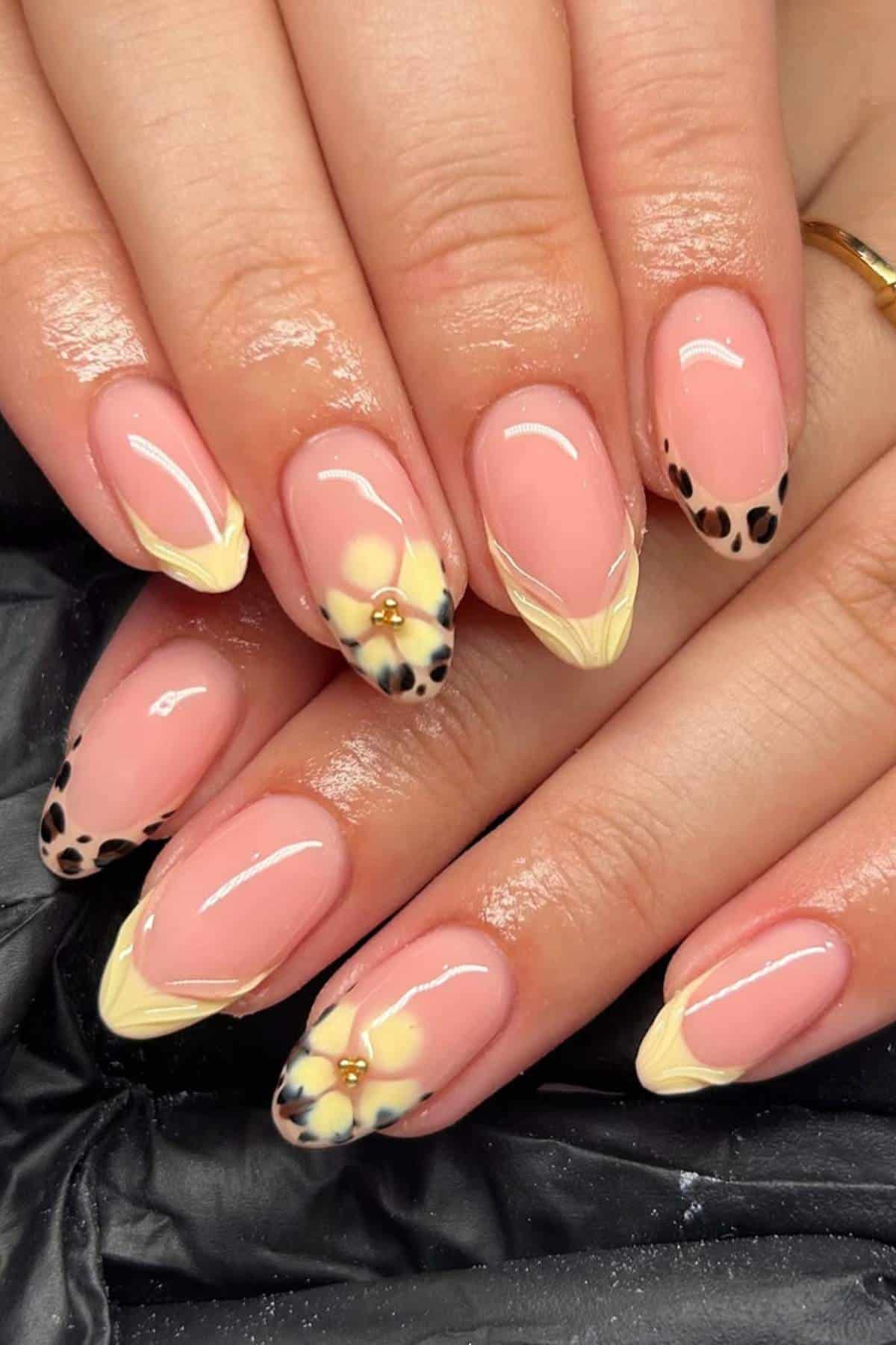 Hands with almond-shaped nails featuring a pink base, yellow French tips, leopard print accents, and small gold embellishments on two nails.