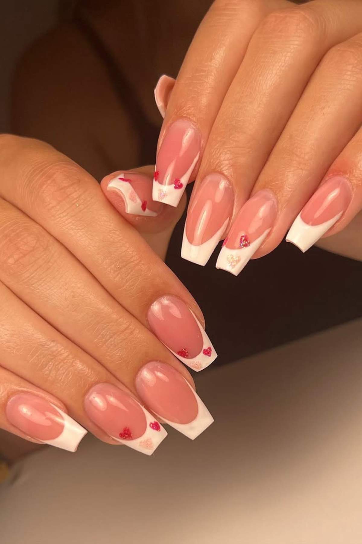 Hands with long, coffin-shaped nails featuring a French tip design, small red and gold heart accents, and one nail with a pink abstract pattern.