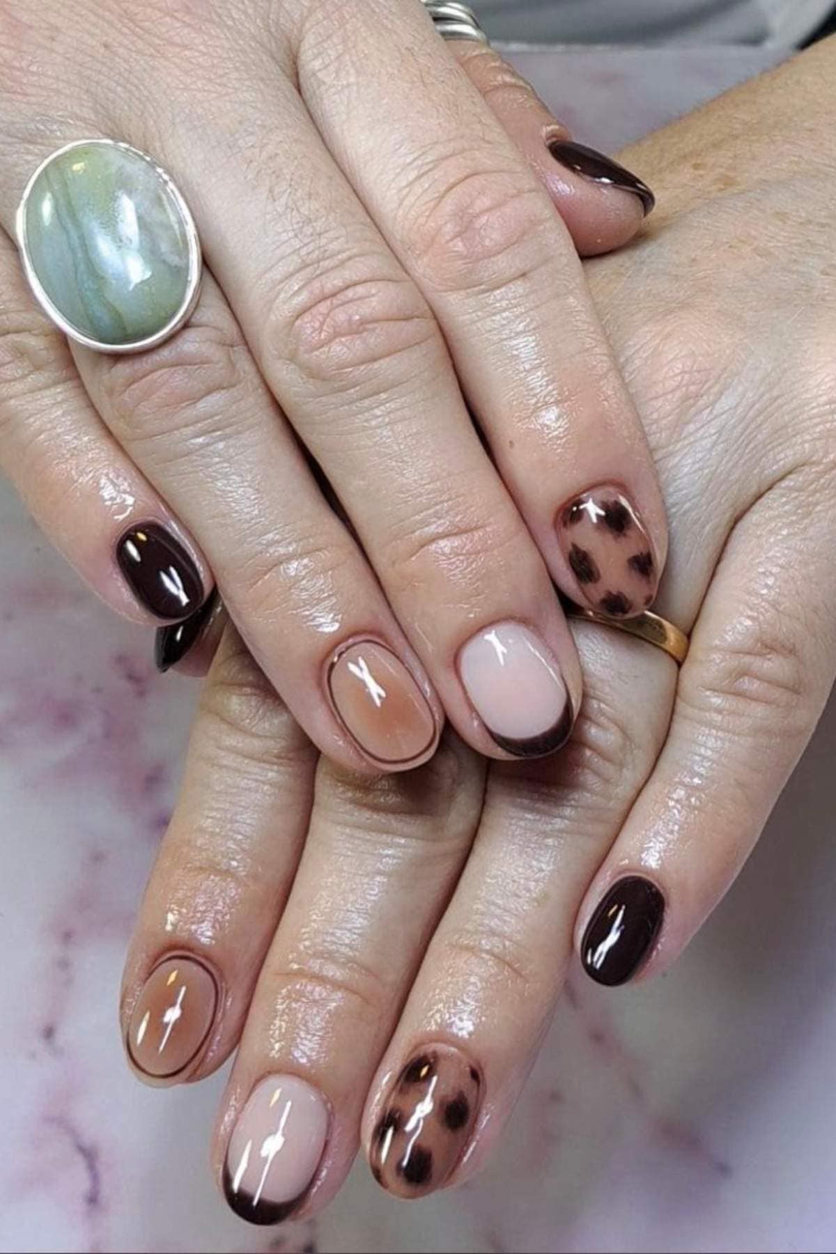 Hands with polished nails featuring a mix of solid dark brown, nude gradient, and brown animal print designs, with a large stone ring on one finger.