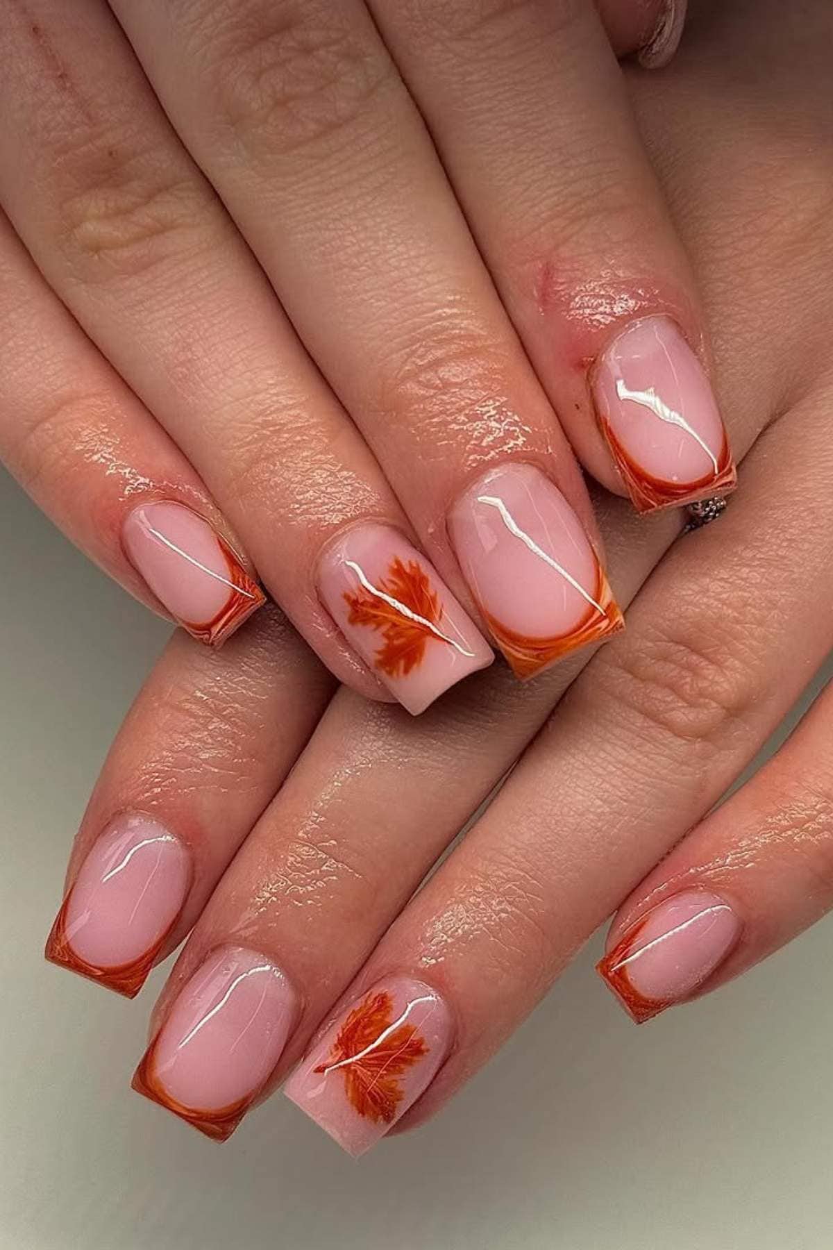 Hands with square, nude-pink acrylic nails featuring orange-tipped French tips and autumn leaf designs on two accent nails.