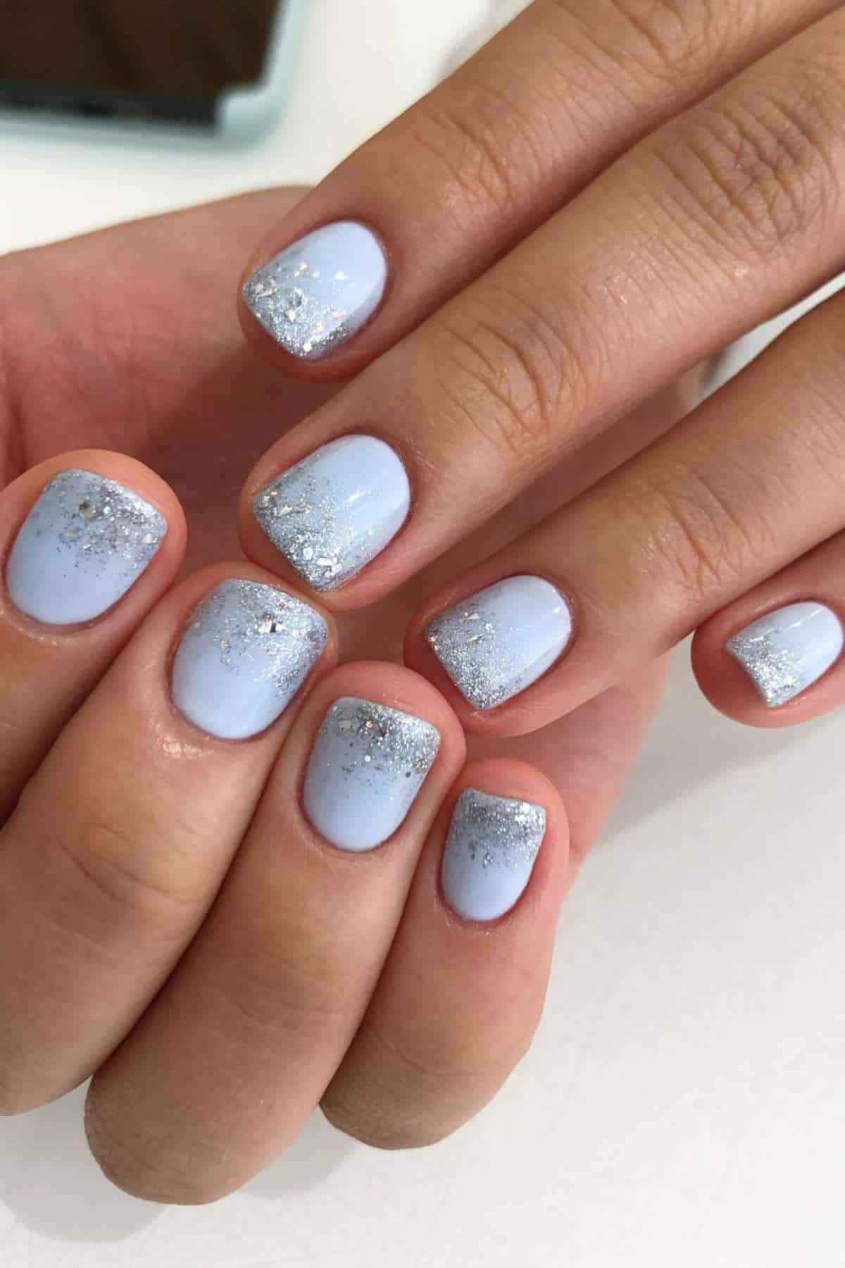 Close-up of hands with short, square-shaped nails painted light blue and decorated with silver glitter at the tips.