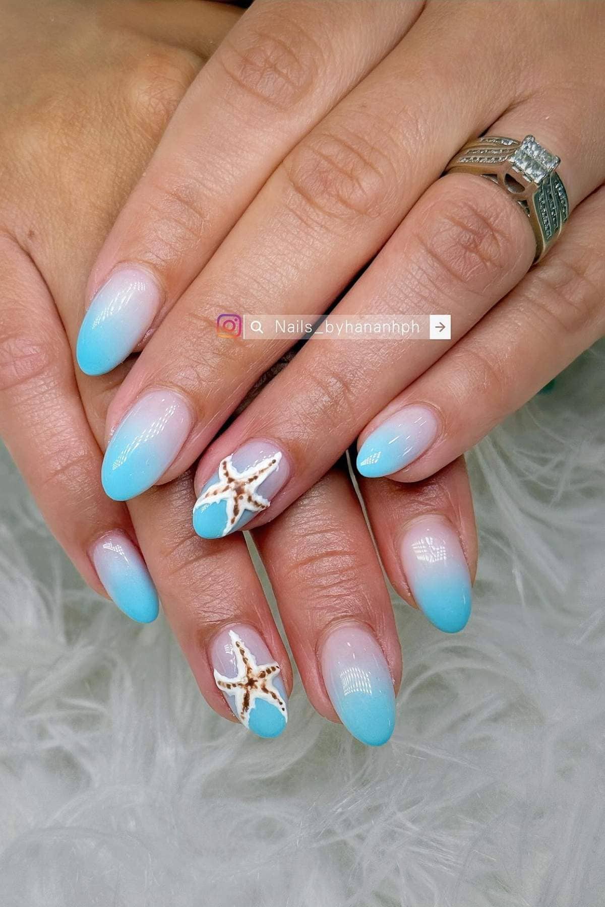 Hands with blue and white ombre manicure; ring fingers feature 3D starfish and seashell nail art. Person wears a silver ring with a large center stone.