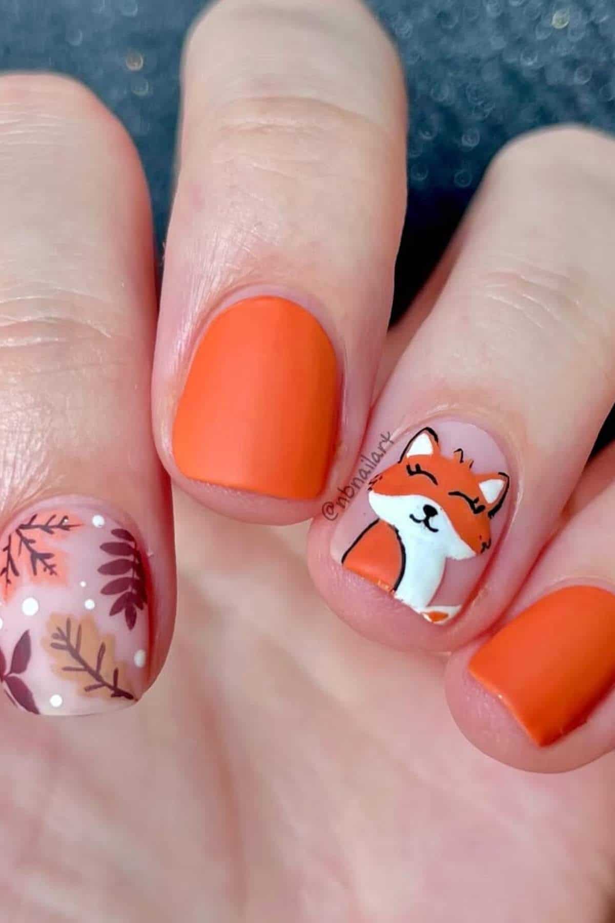 Hand with orange nail polish, one nail featuring a painted fox face, and another nail decorated with leaf patterns and small white dots.
