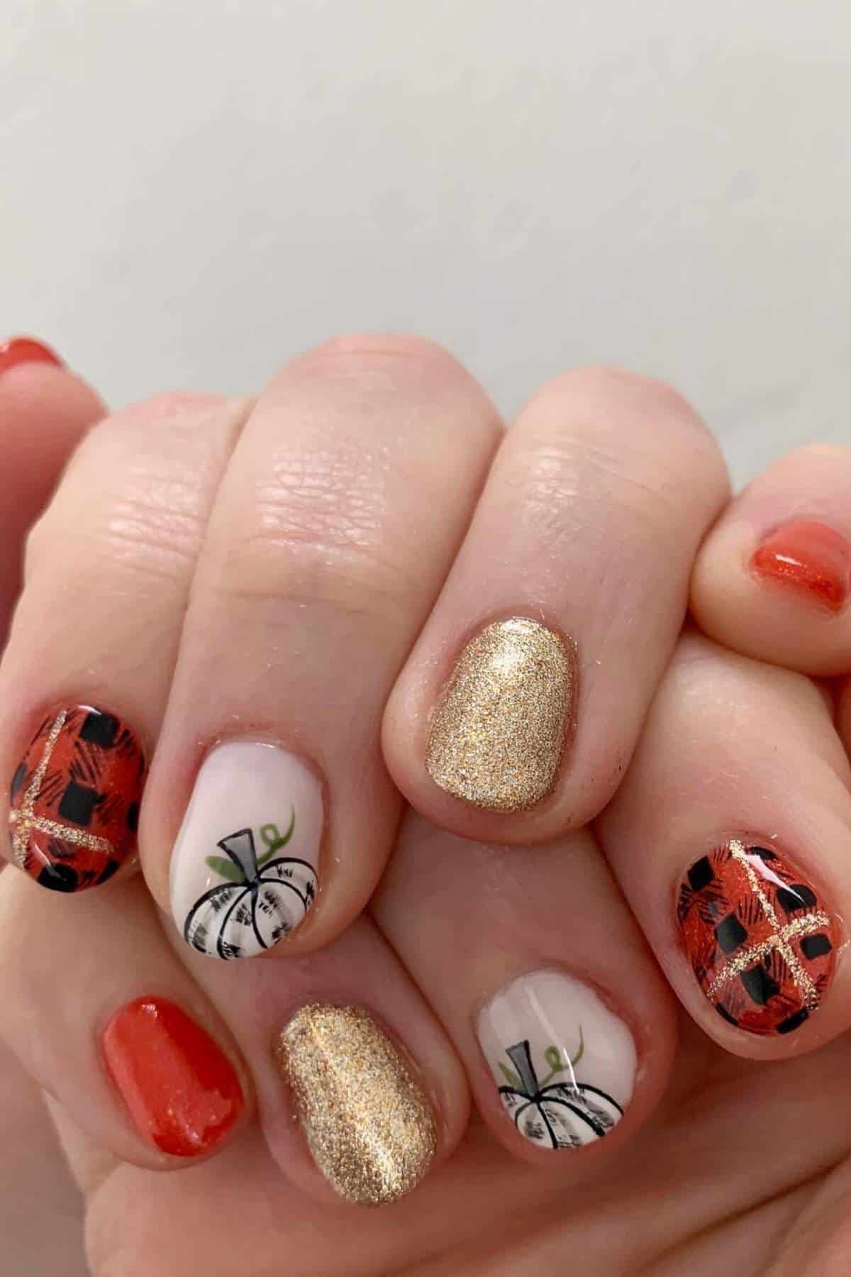 Close-up of a hand with fall-themed nail art featuring red, gold glitter, plaid patterns, and white pumpkin designs.