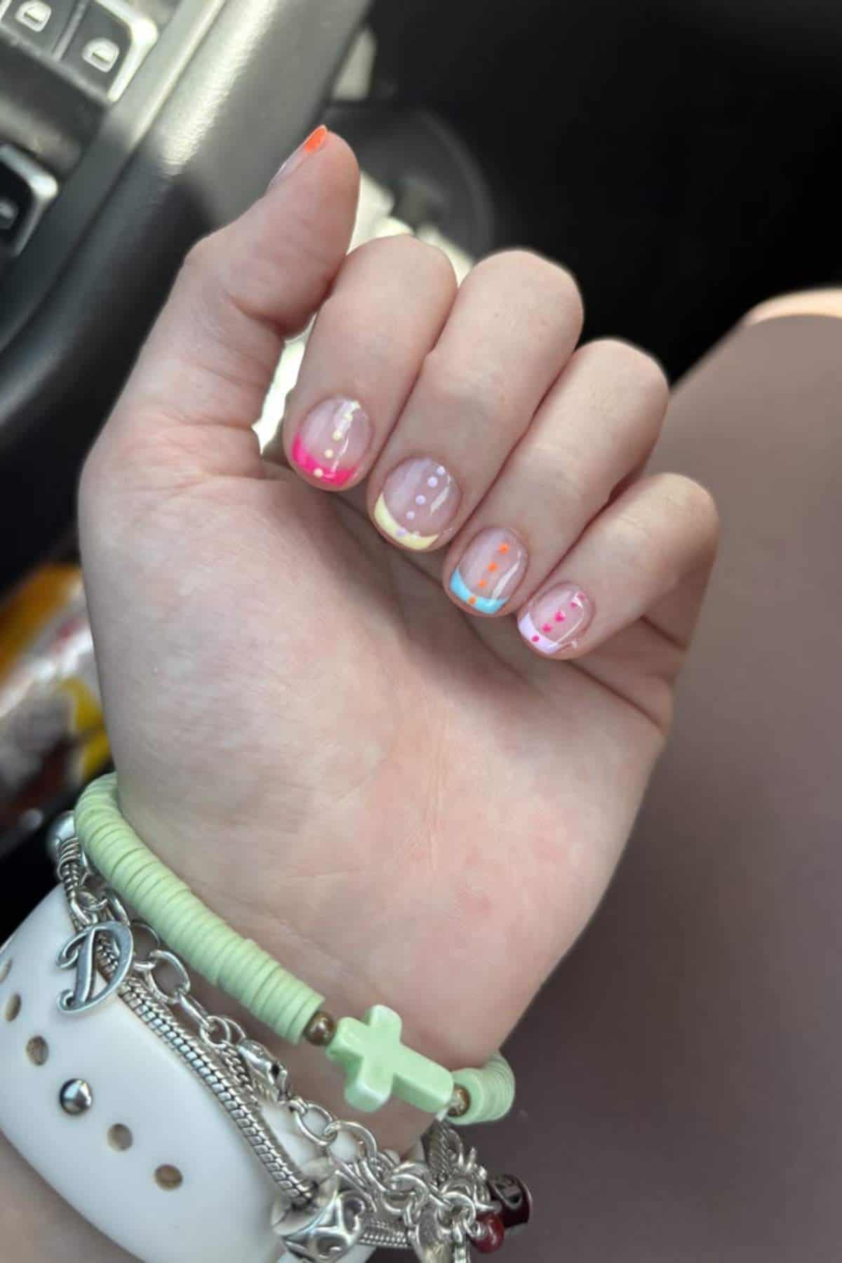A hand with painted nails featuring multicolored French tips and dots, wearing silver bracelets and a green bead bracelet, resting on a lap in a car.