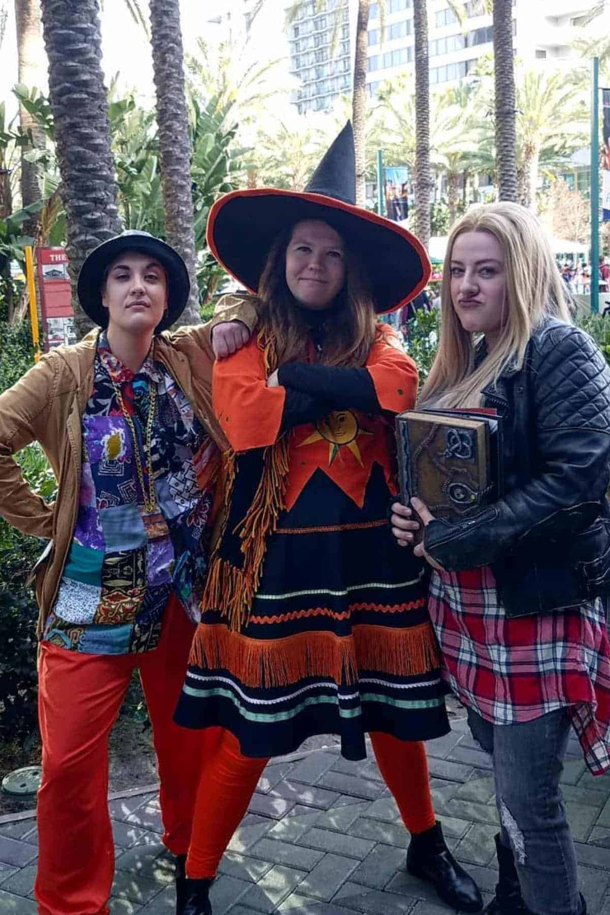 Three people in colorful costumes pose outdoors; one wears a witch hat and dress, another holds a book prop, and the third wears a patterned shirt and bucket hat. Palm trees are in the background.
