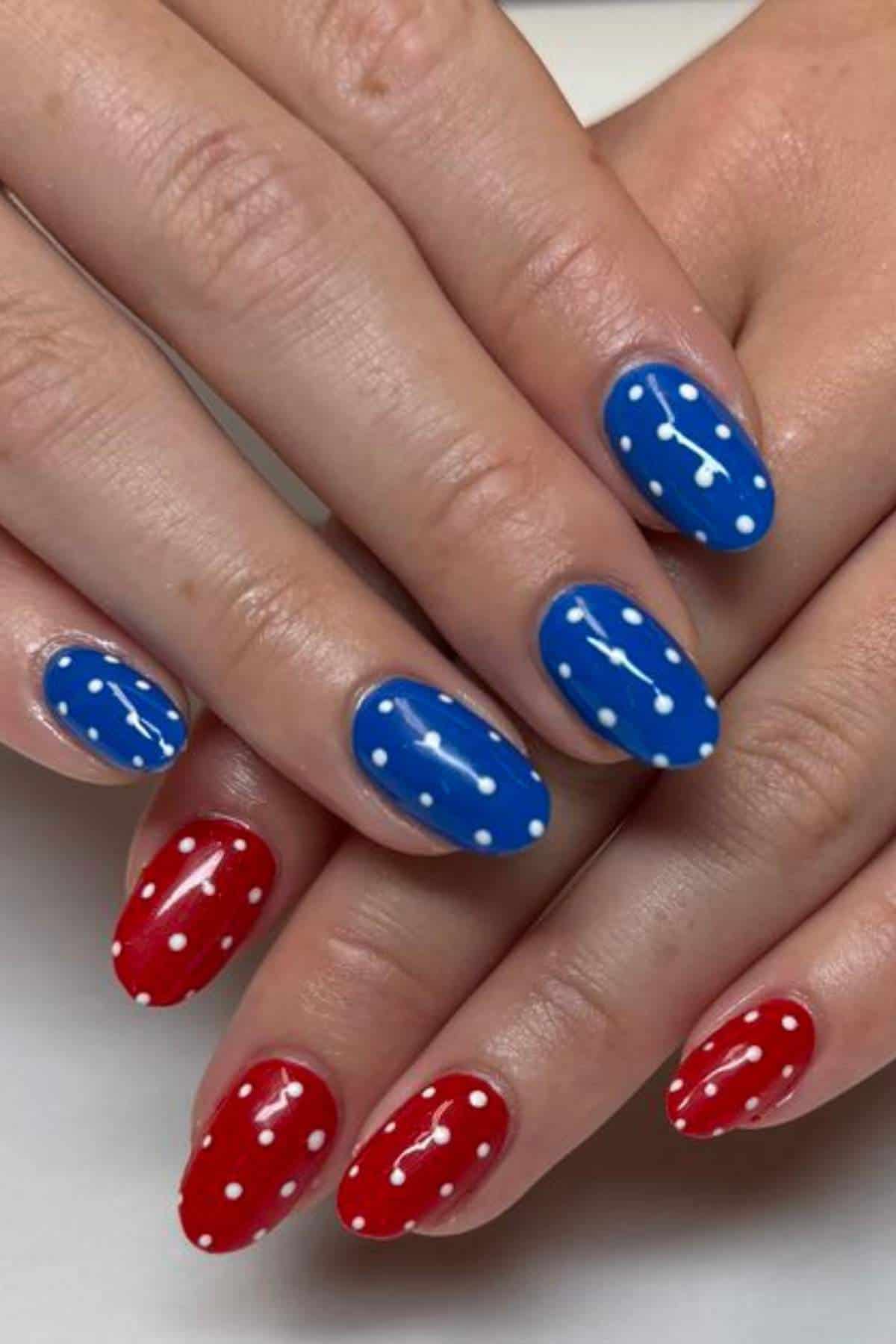 Hands with manicured nails painted red and blue, each nail decorated with white polka dots arranged in lines and around the edges.