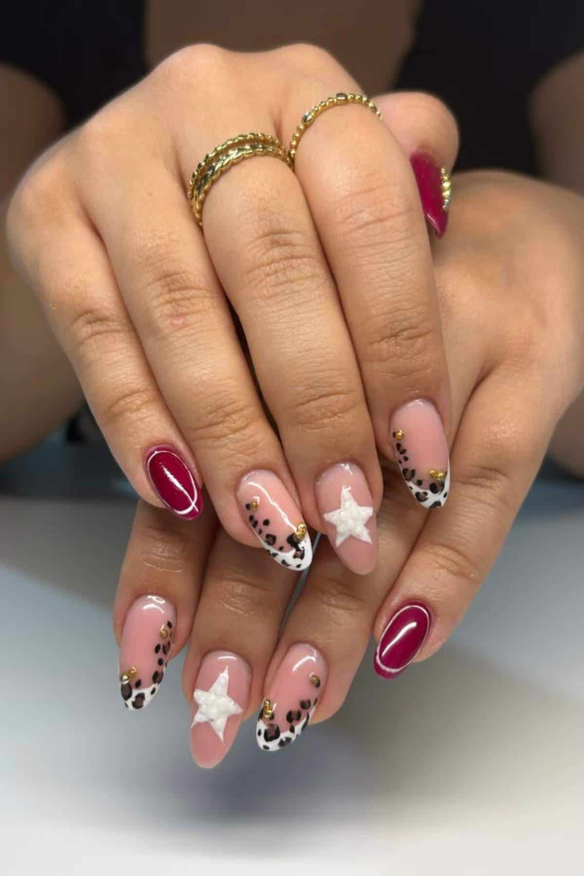 Hands with almond-shaped nails featuring red polish, cheetah print tips, gold studs, and white 3D star accents. The person wears gold rings on several fingers.