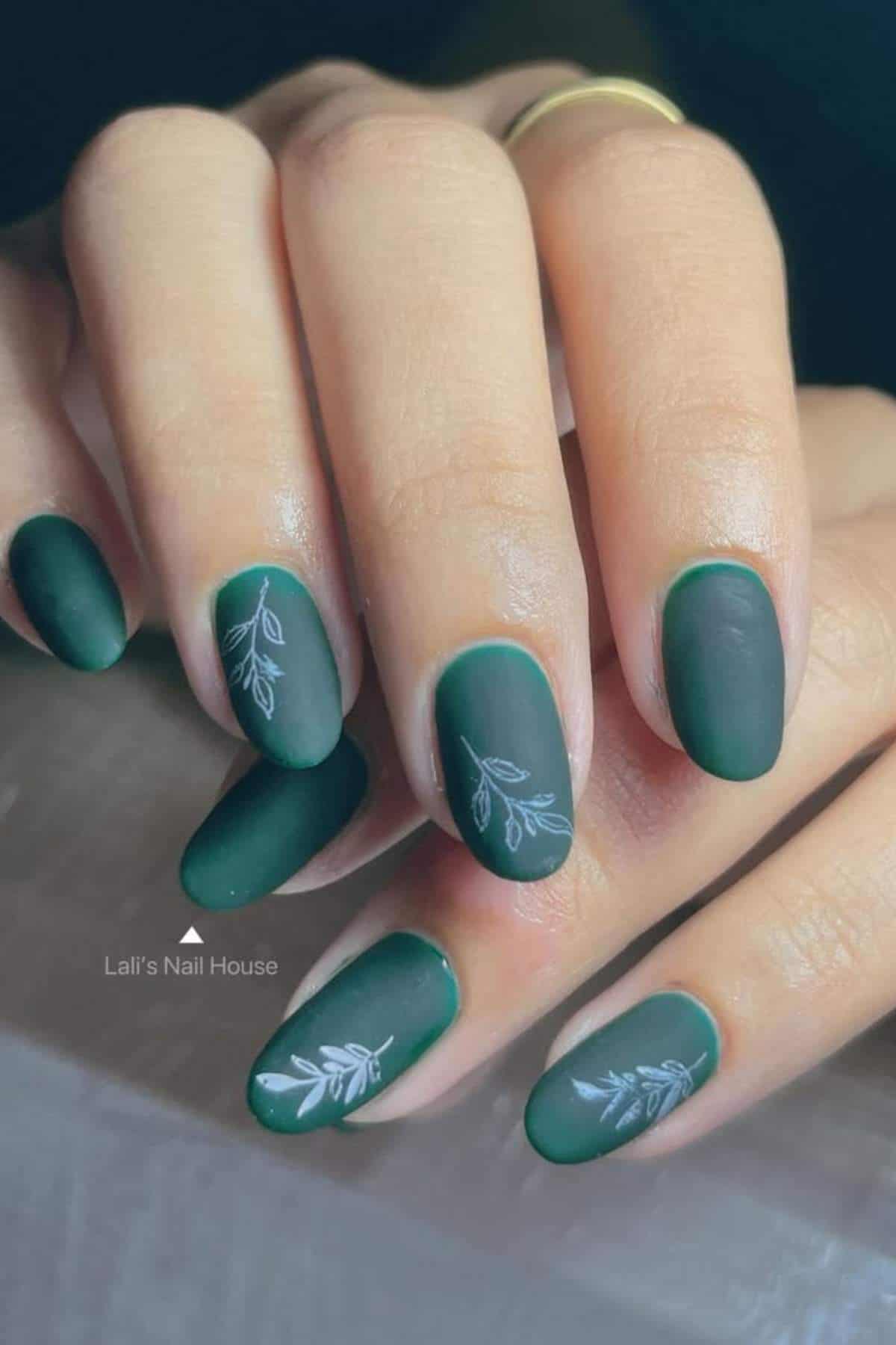 A hand with matte dark green almond-shaped nails featuring white leafy branch designs on two nails.