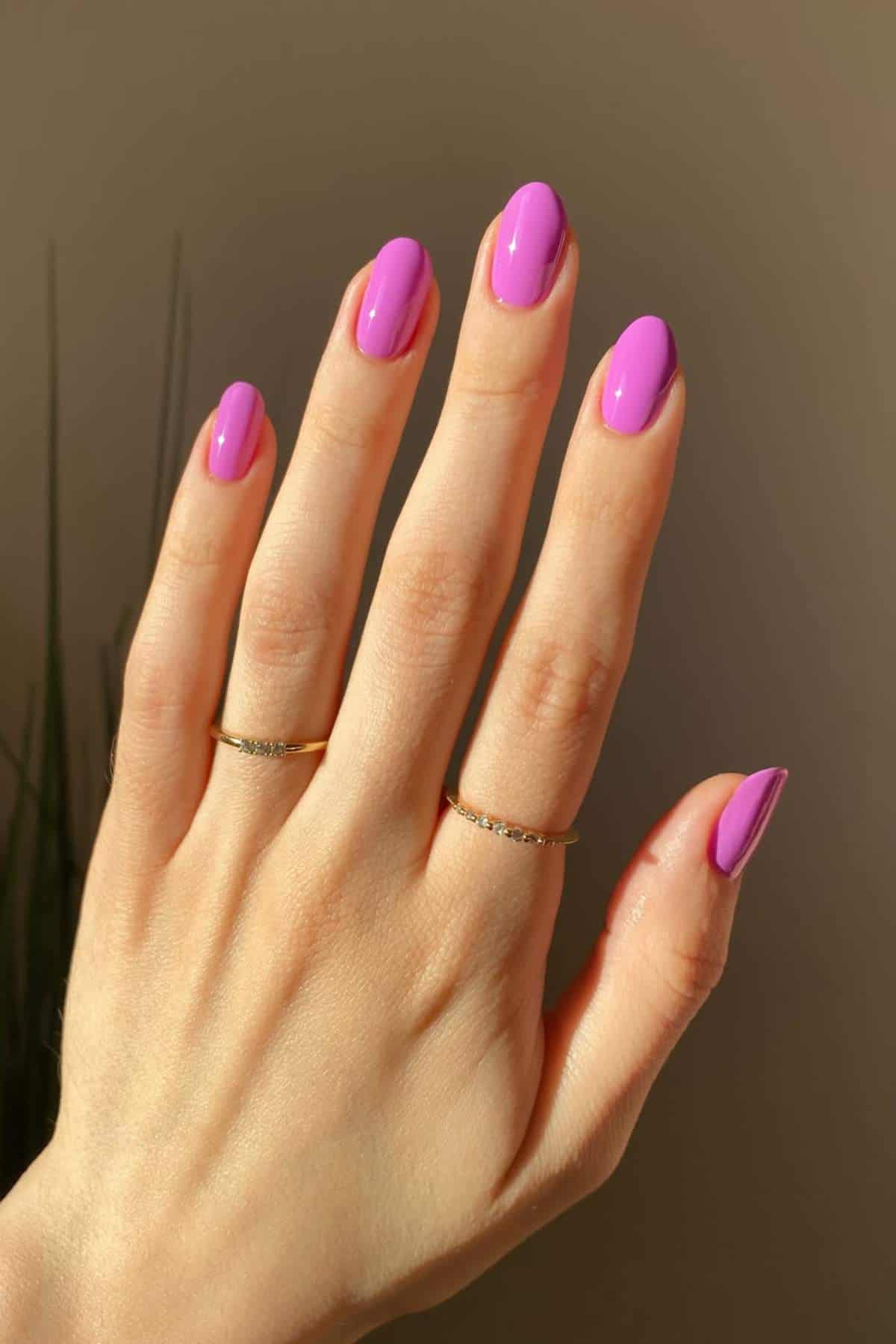 A hand with neatly manicured nails painted bright pink, wearing two thin gold rings on the middle and ring fingers, against a neutral background.