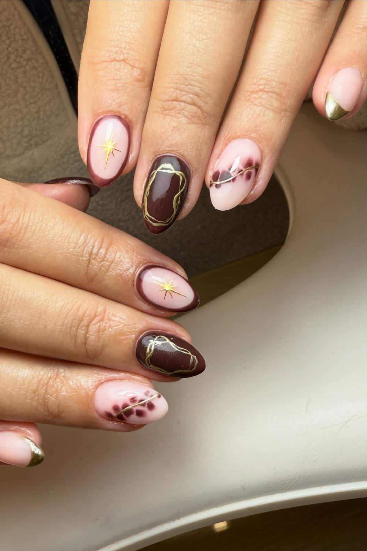 Hands with almond-shaped nails featuring brown and pink polish, abstract gold lines, small star designs, and gold-tipped accents.