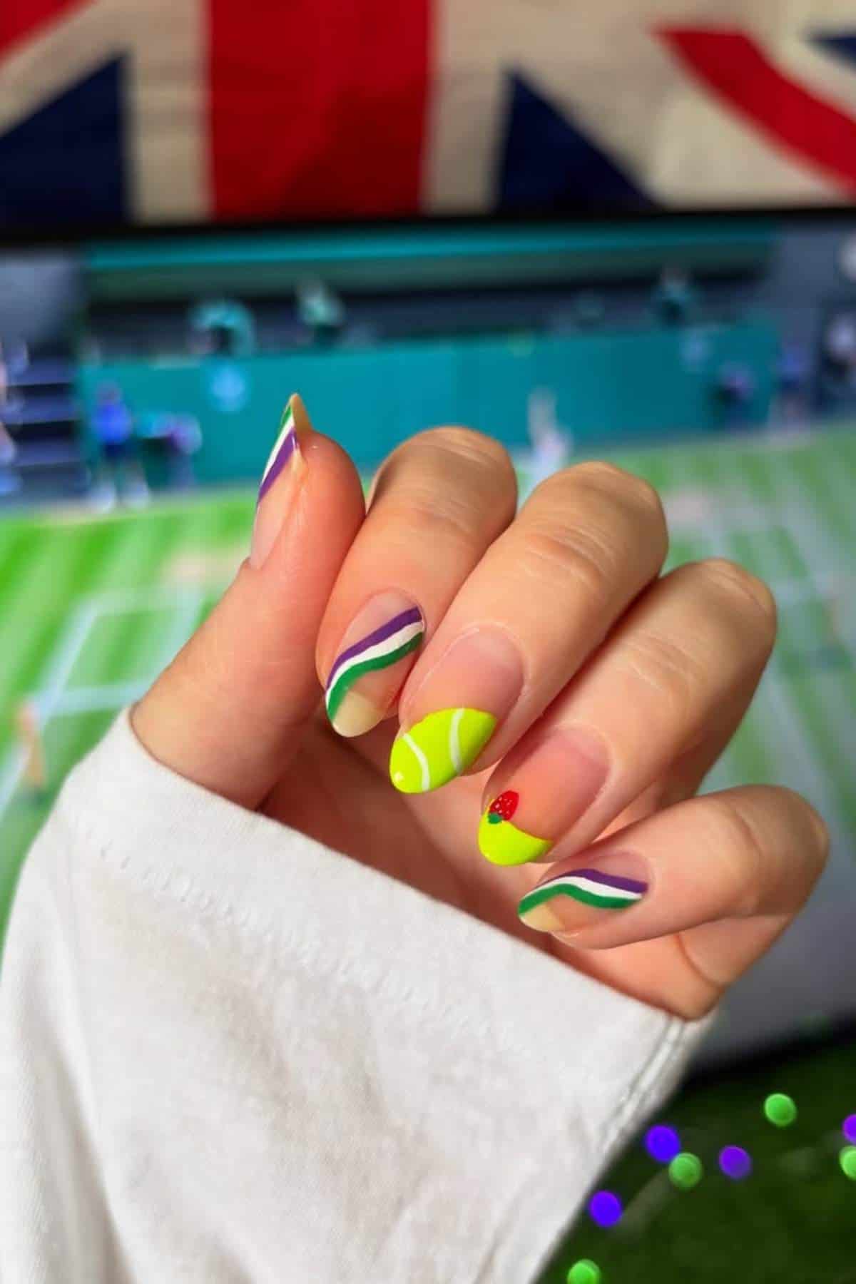 A hand with multicolored abstract nail art, including a tennis ball design on one nail, is held up in front of a TV showing a tennis match and a British flag.