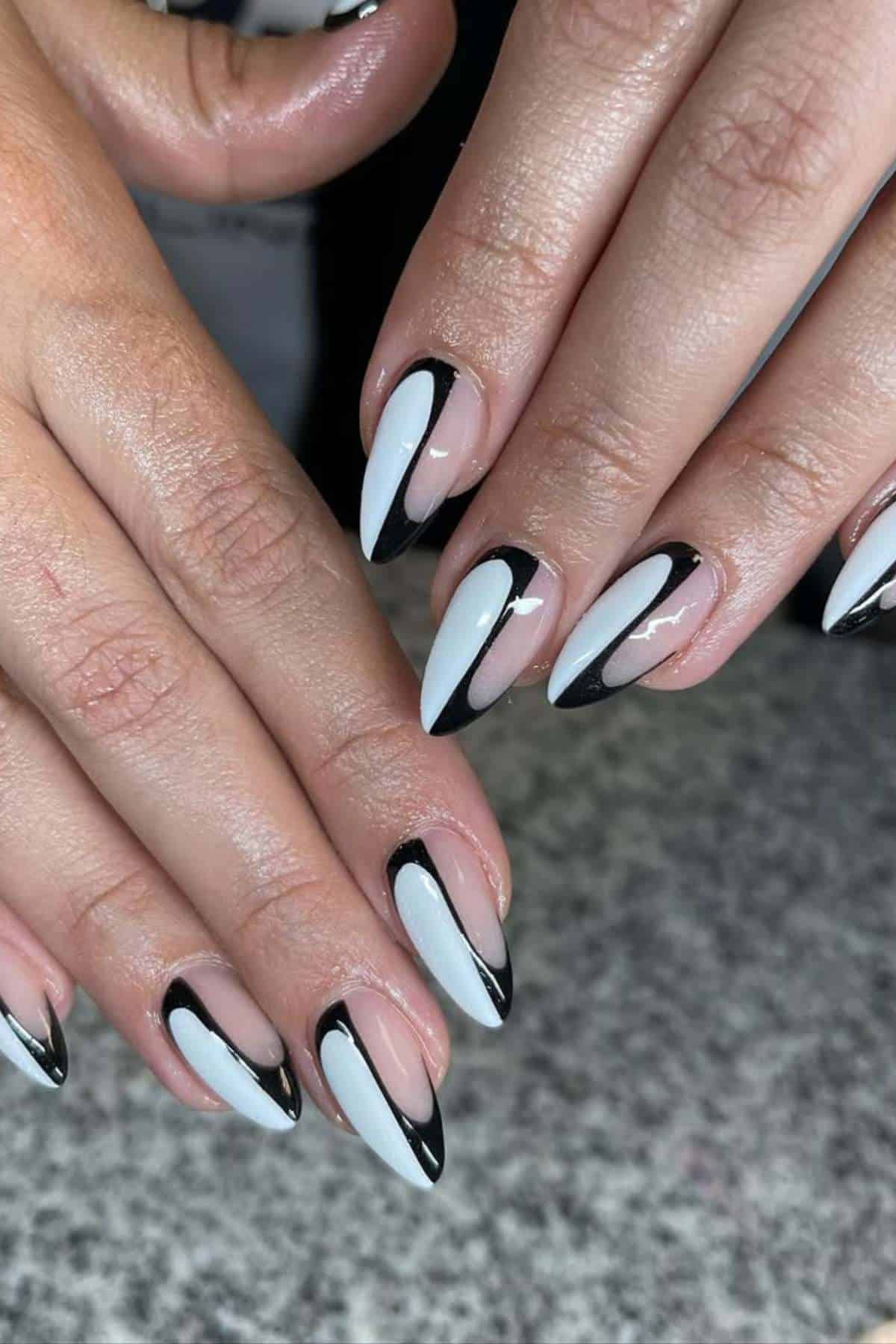Hands with almond-shaped nails featuring a white base and black outlined nail art, displayed on a speckled gray surface.