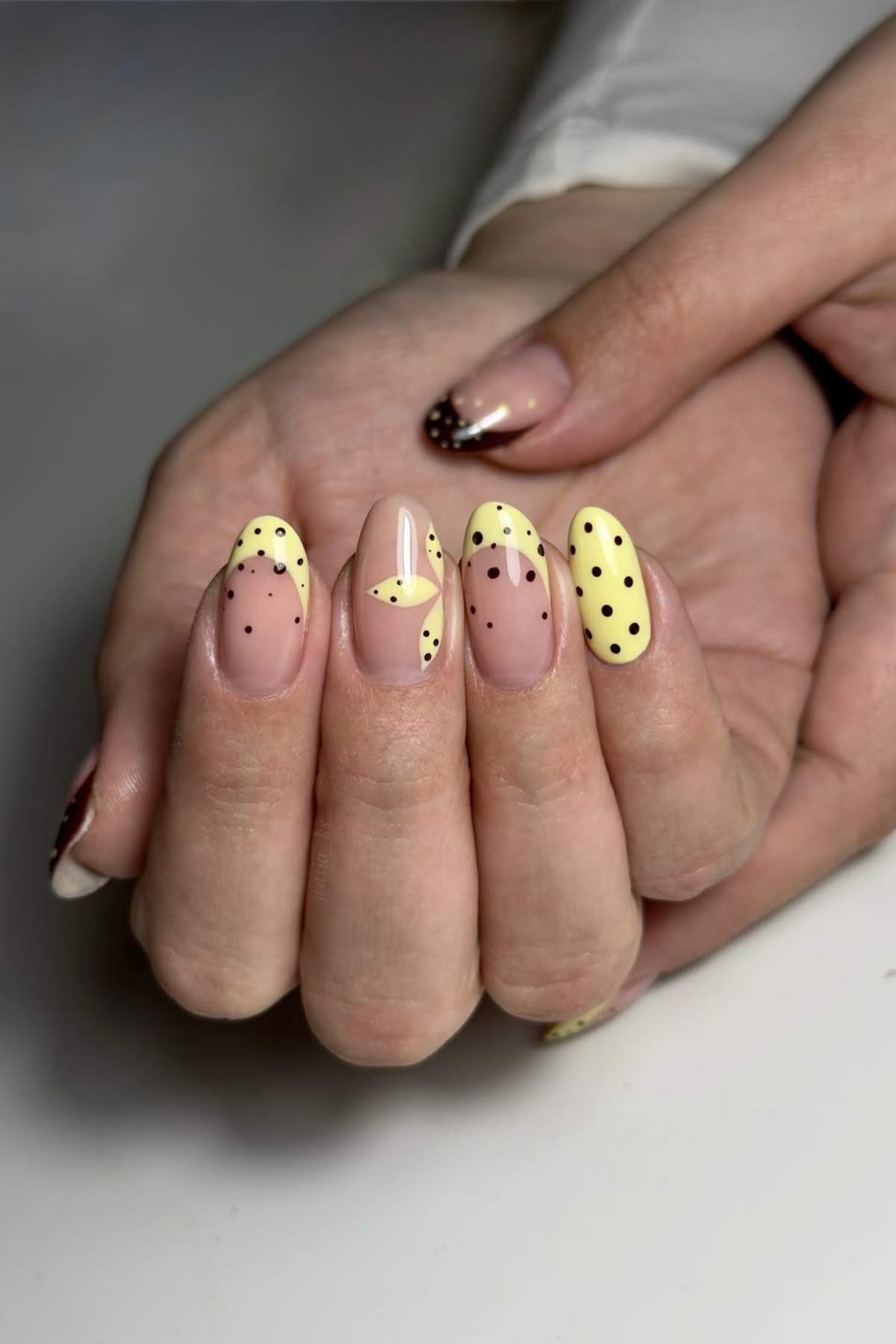 A hand with almond-shaped nails featuring yellow and nude polish, black polka dots, and a butterfly design on the middle finger.