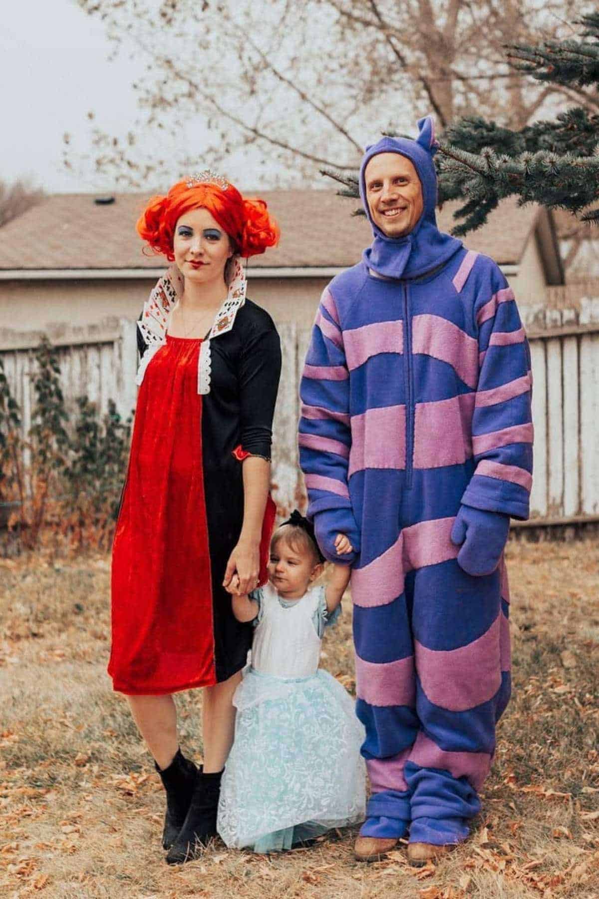 Three people in costumes stand outside: an adult in a red and black dress with orange hair, a child in a light blue dress, and an adult in a purple and pink striped suit with a tail and ears.