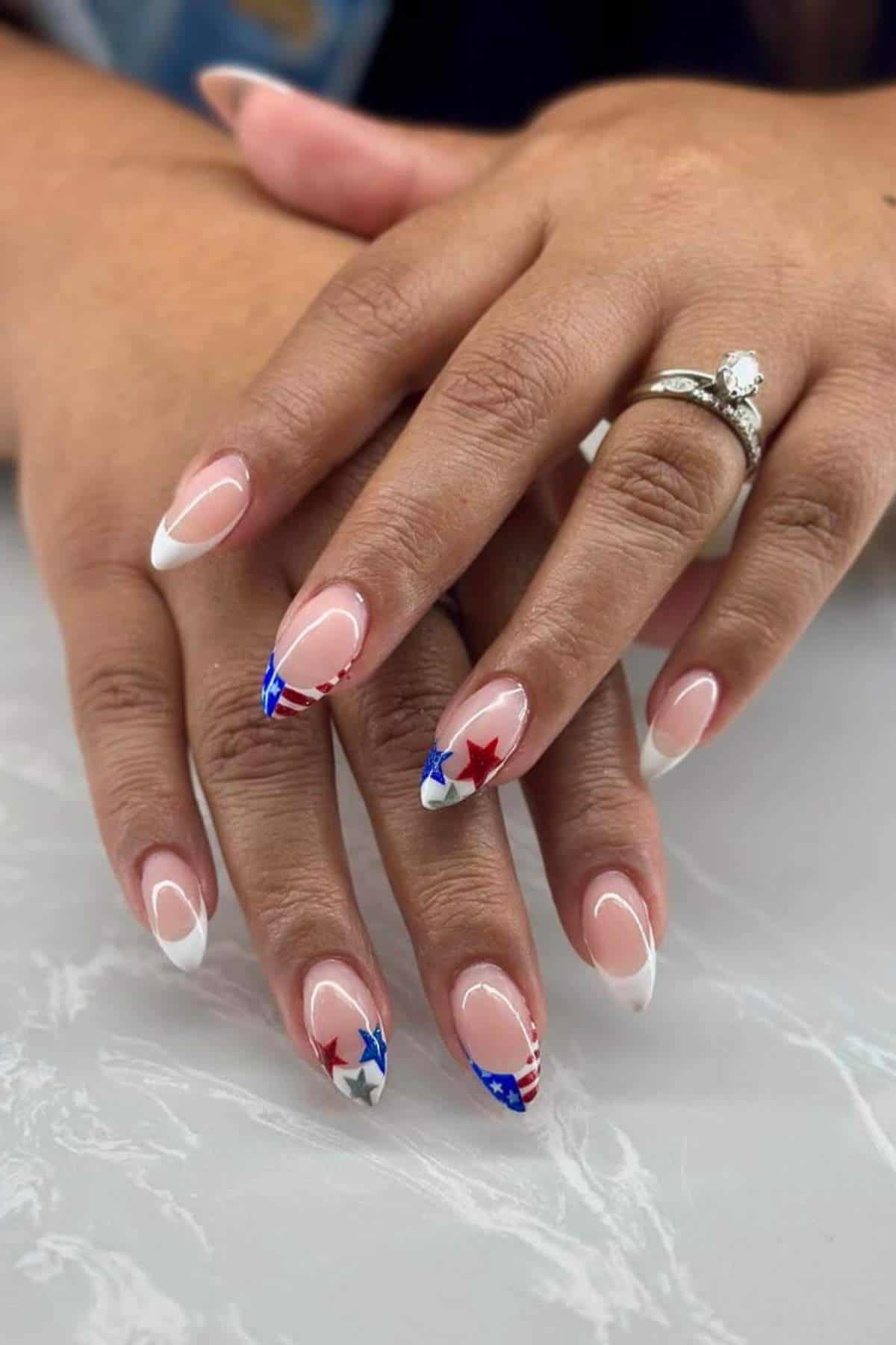Hands with almond-shaped nails featuring white French tips and patriotic designs with red and blue stars and stripes on a few accent nails, resting on a light surface.