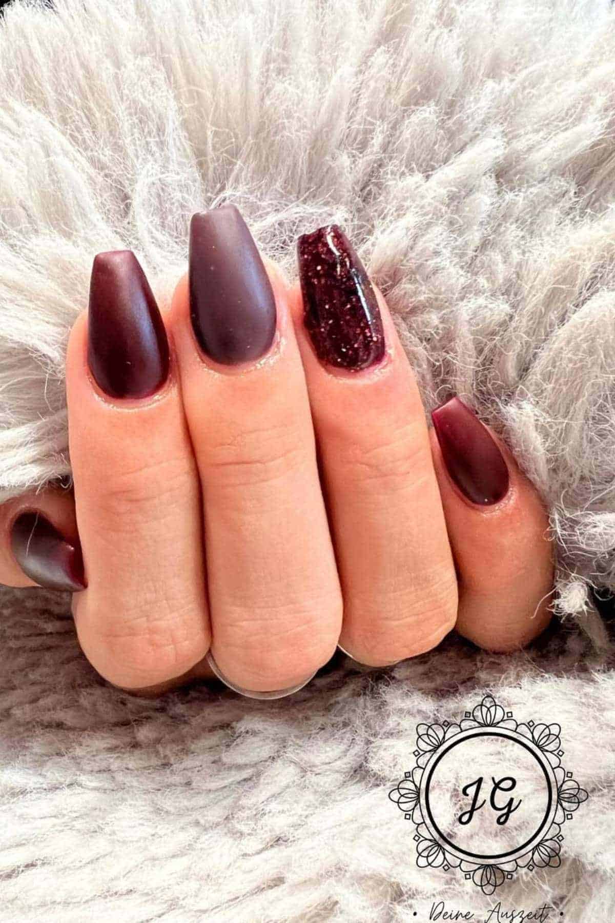 A hand with matte burgundy coffin-shaped nails, with one accent nail featuring glitter, resting on a soft, gray textured fabric.