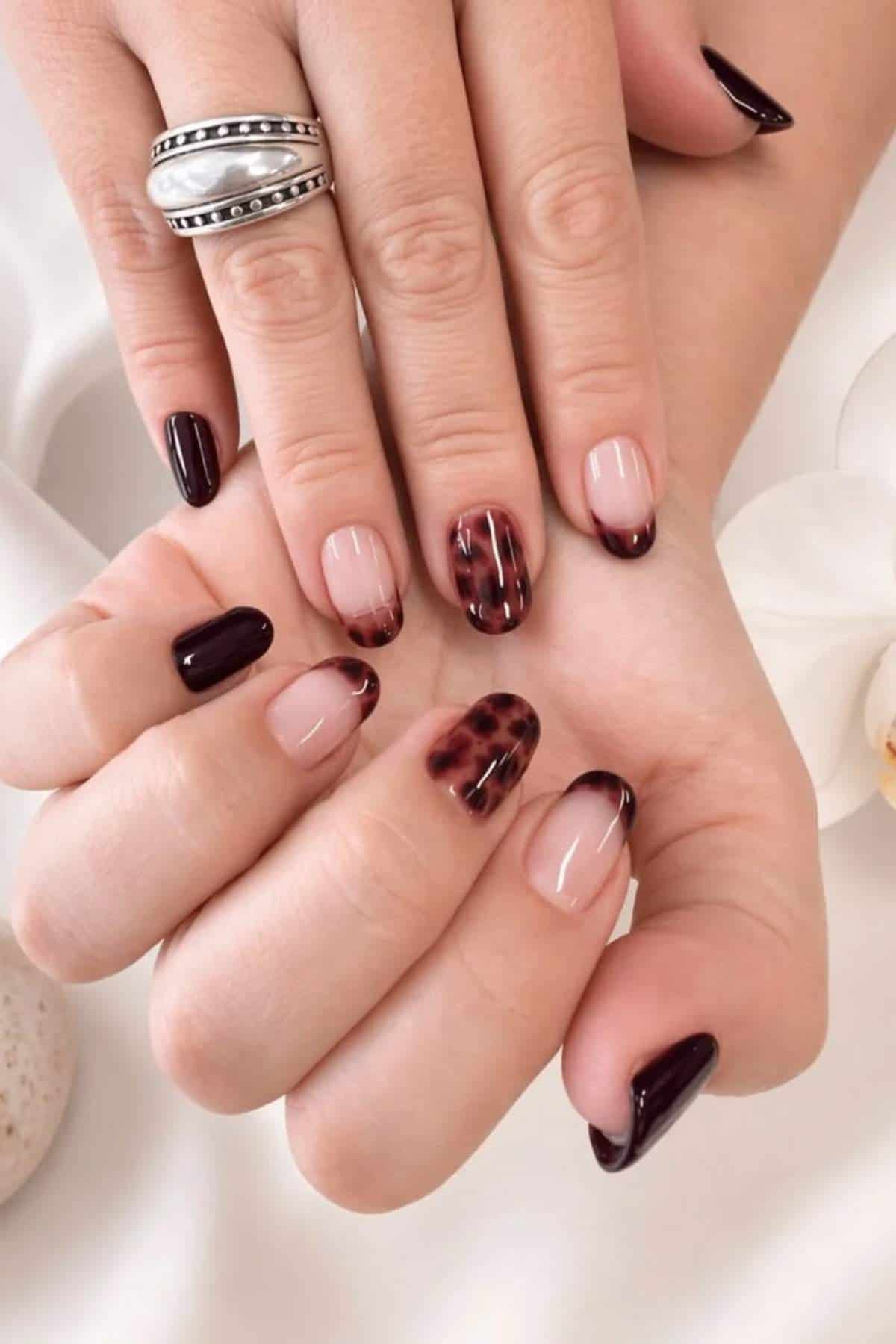 Hands with dark maroon and tortoiseshell French tip nail art, one finger wears a silver ring with black detailing.