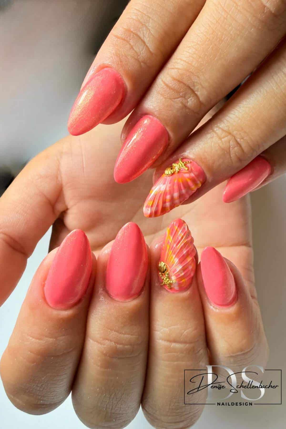 A hand with almond-shaped, coral pink nails; two accent nails feature shell-like designs and gold embellishments.