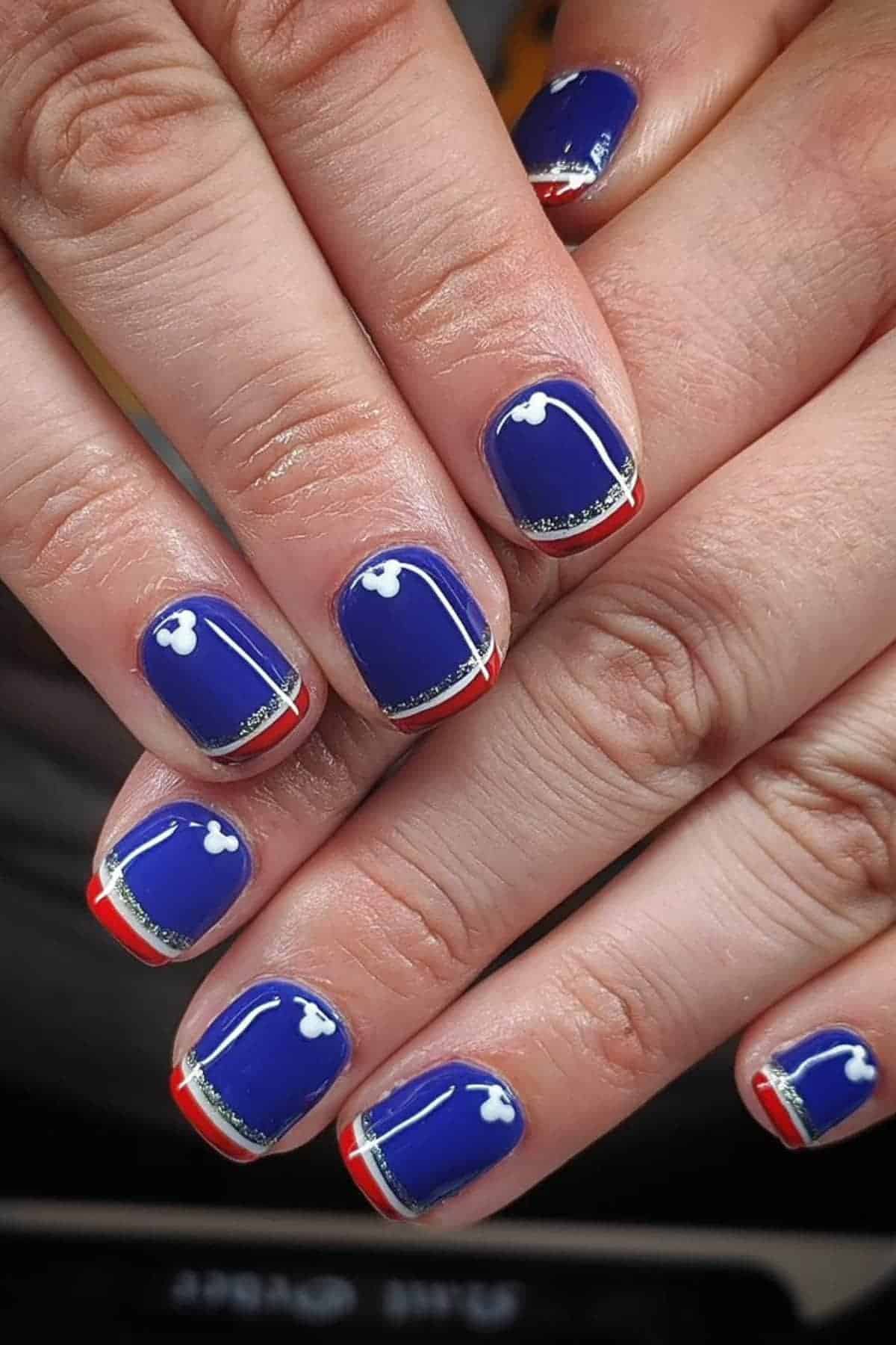 Close-up of hands with blue nail polish, white Mickey Mouse head designs, silver glitter stripe, and red and white tips on short, square-shaped nails.