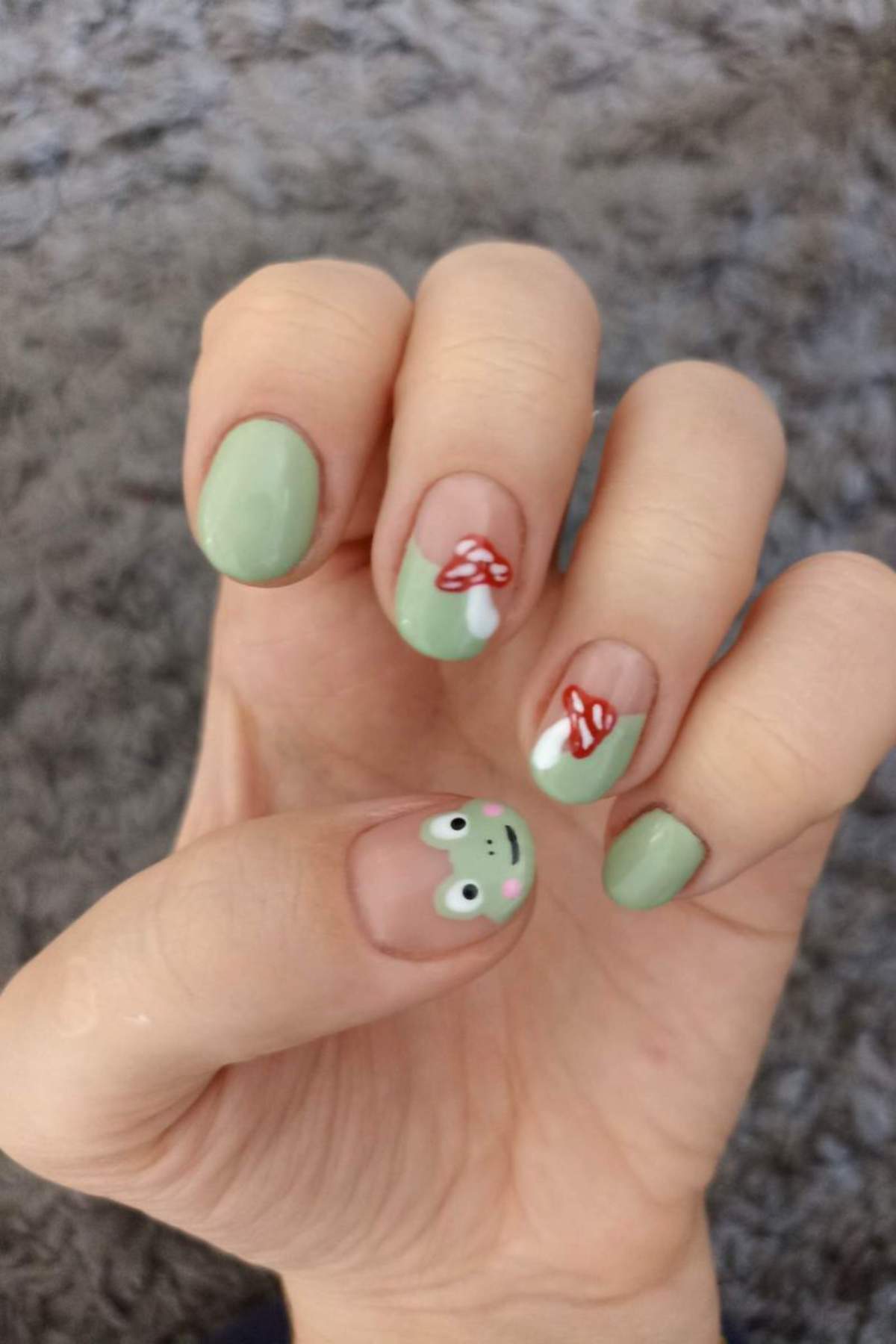 A hand with light green nail polish, featuring a frog face design on the thumb and red mushroom designs on two fingers, against a gray textured background.
