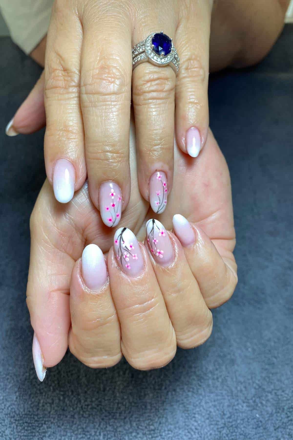 A woman's hands display an almond-shaped manicure featuring a pink and white gradient with delicate cherry blossom nail art. A large sapphire ring adorns her index finger.