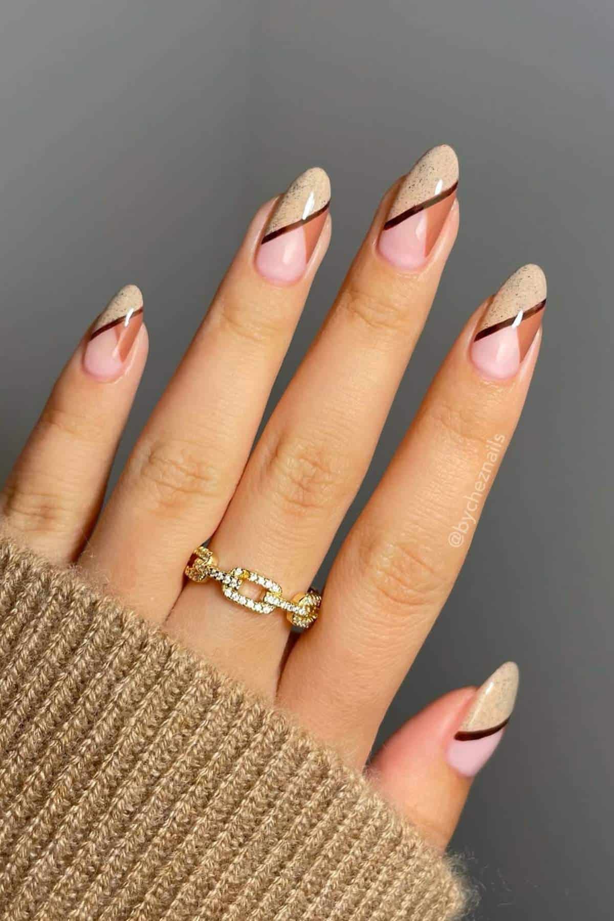 A hand with almond-shaped nails featuring a nude and beige design with diagonal brown lines. The person is wearing a gold ring and a beige sweater.