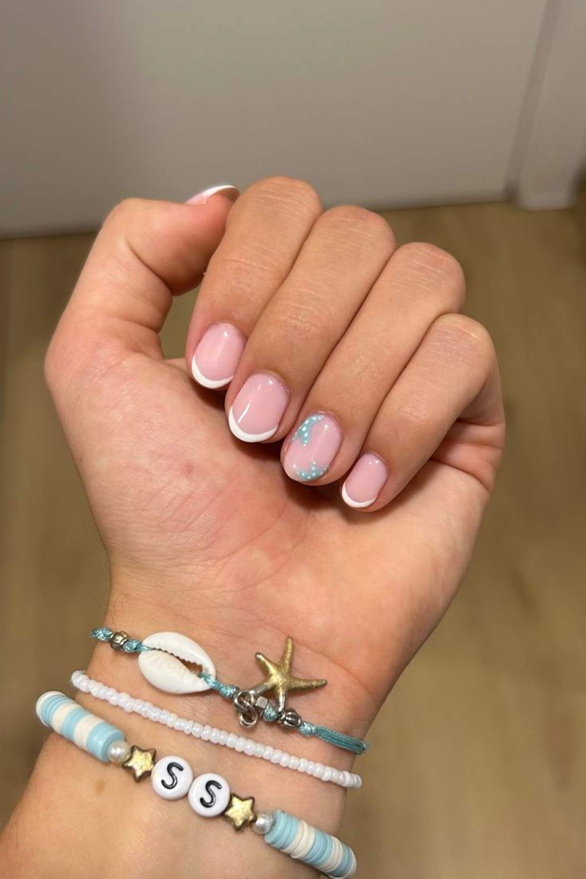 A hand with a French manicure featuring a blue glitter accent on one nail, wearing three beaded bracelets with a shell, starfish charm, and letter beads.